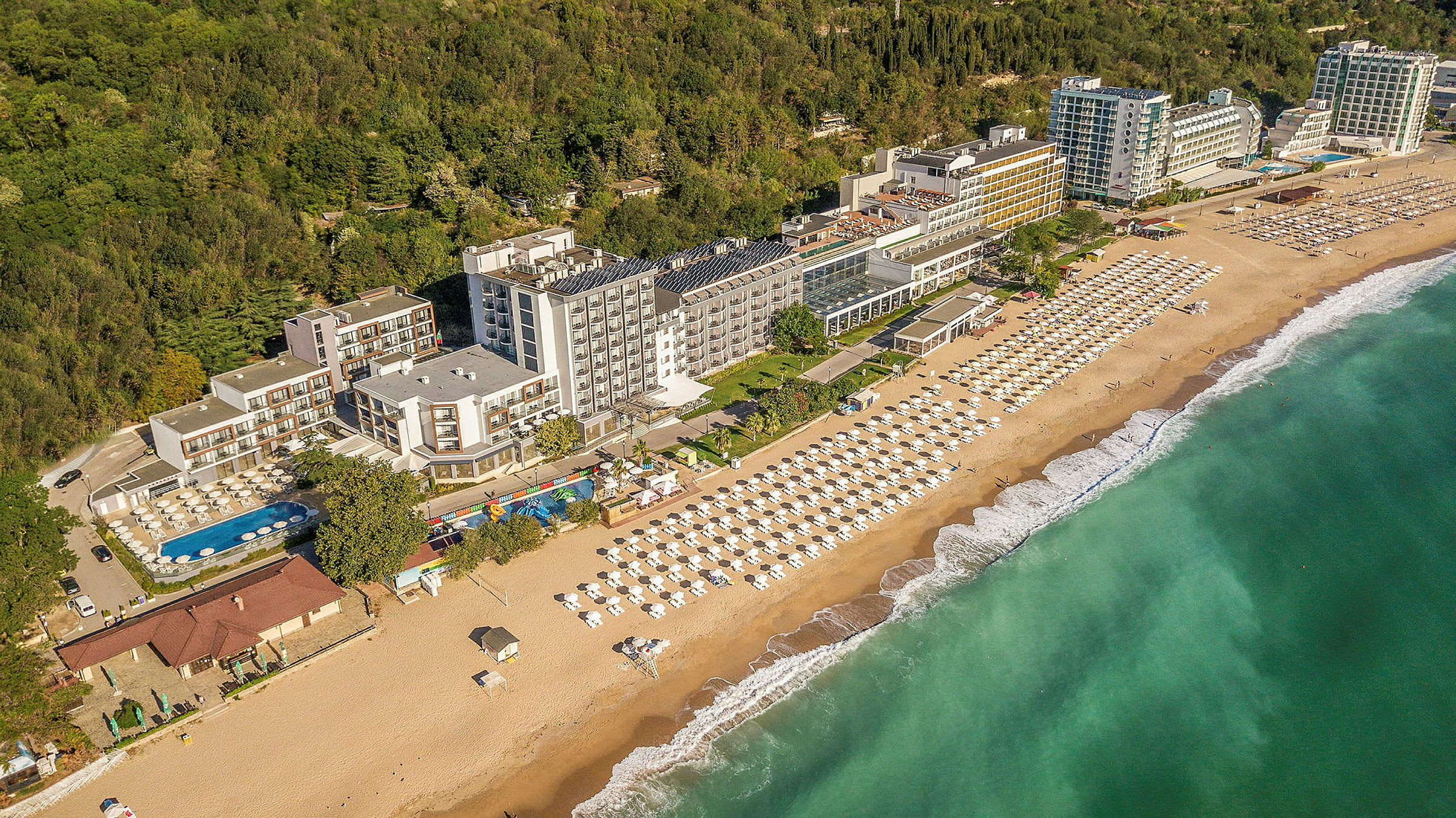 Udsigt over hotel ved kysten med strand og parasoller samt grønne områder i baggrunden. (AI-genereret tekst)