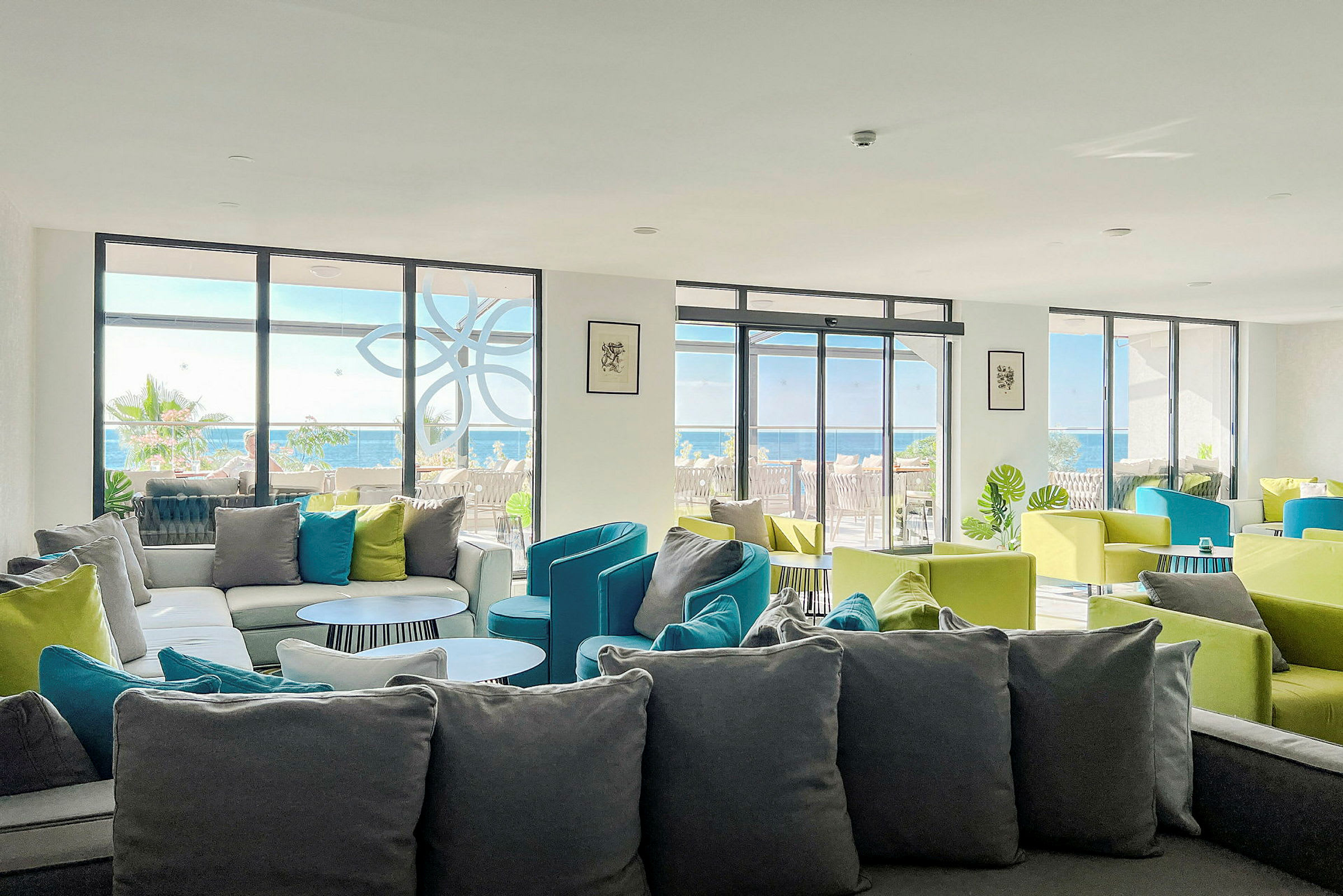 En lounge med sofaer og farverige stole, og vinduer vendende mod en terrasse og havet. (AI-genereret tekst)
