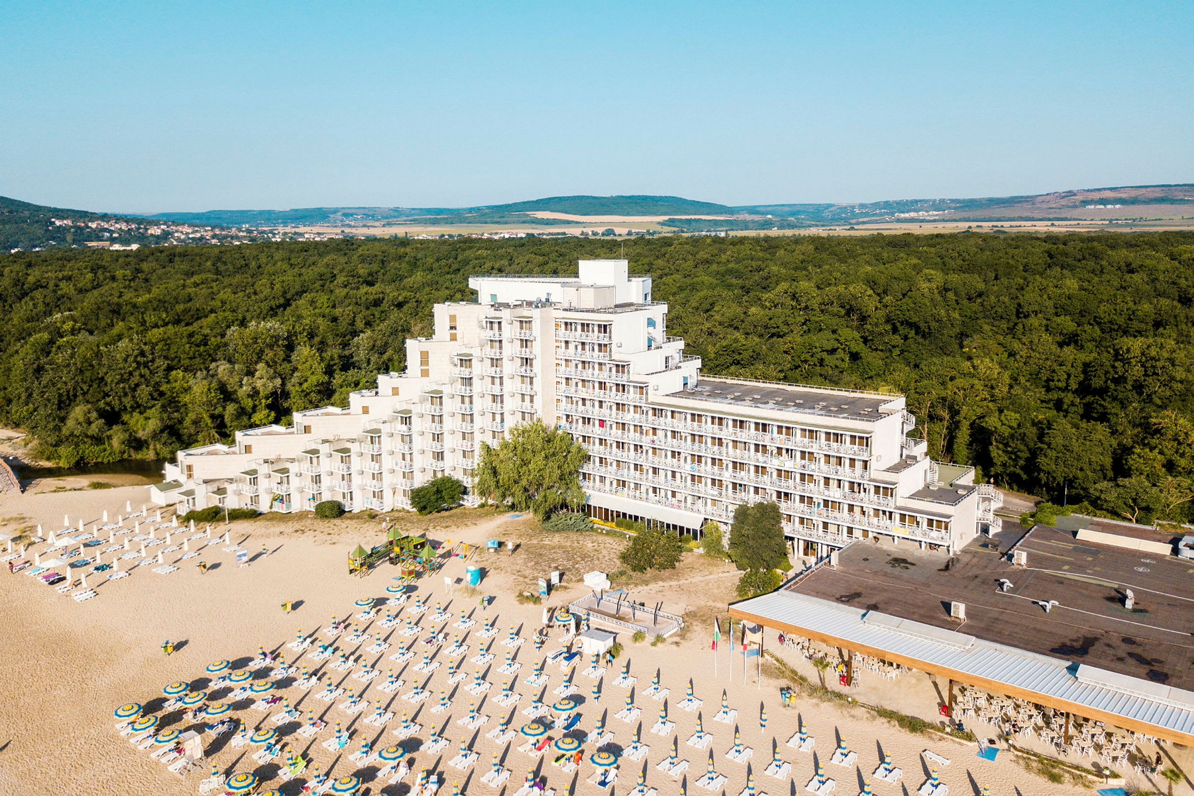 Billede af et hotel ved en strand med liggestole og parasoller, omgivet af skov. (AI-genereret tekst)