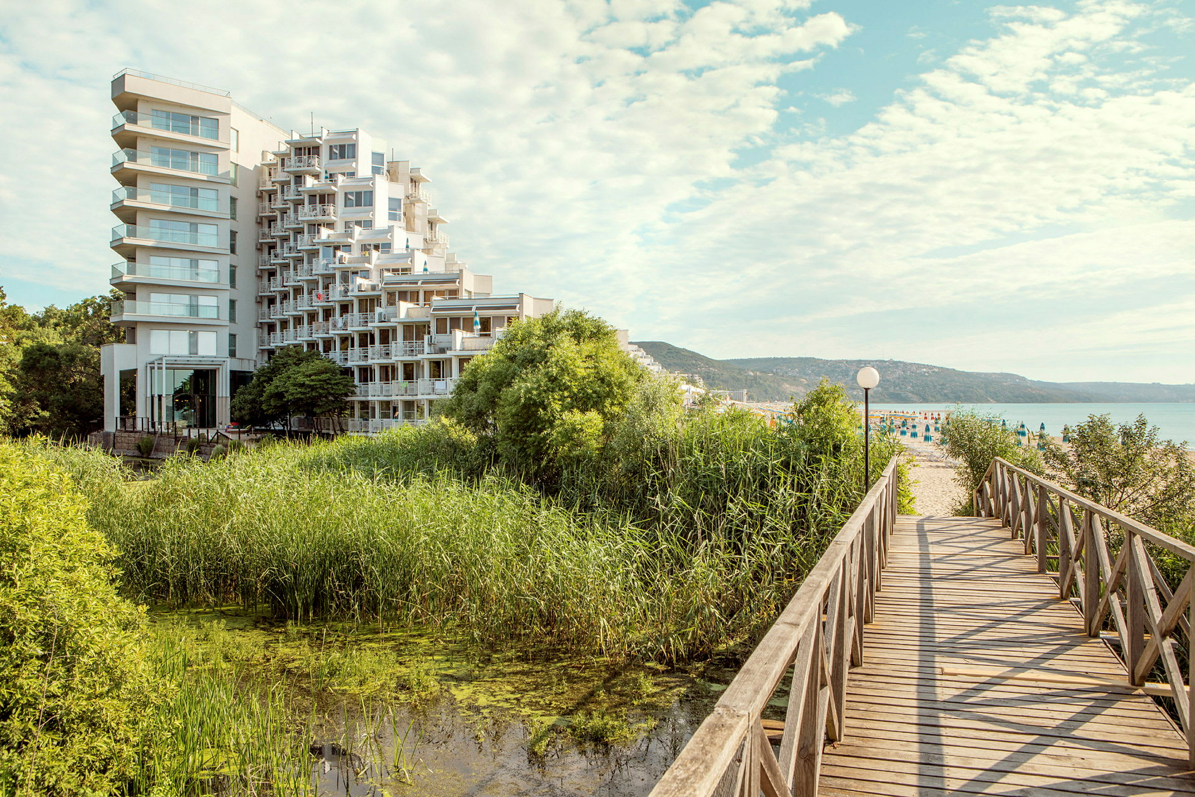 En moderne hotelbygning ligger tæt på et område med grønt og vand, og en træbro fører mod en sandstrand. (AI-genereret tekst)