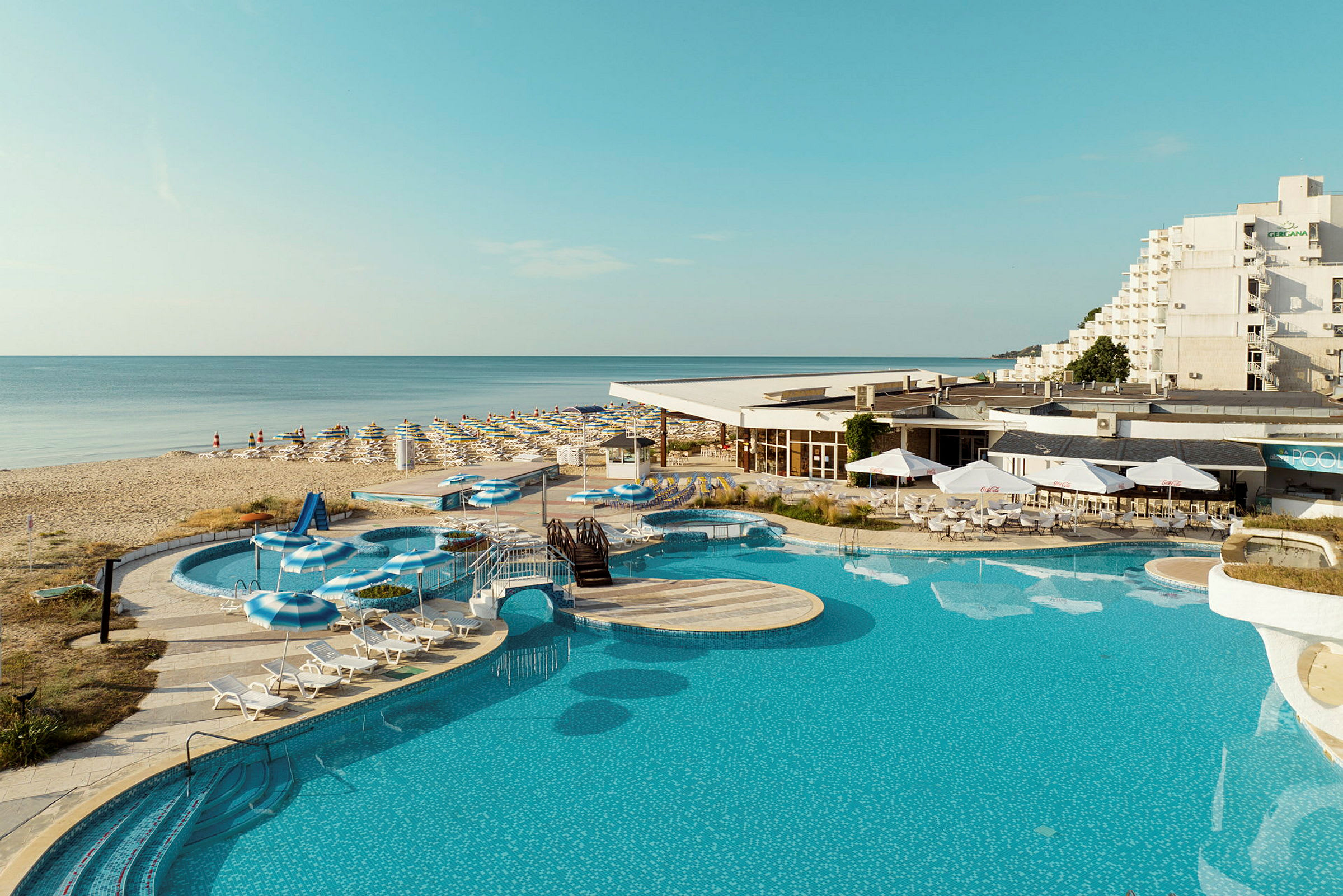Et billede af en pool, liggestole og parasoller ved et hotel tæt på en sandstrand. (AI-genereret tekst)