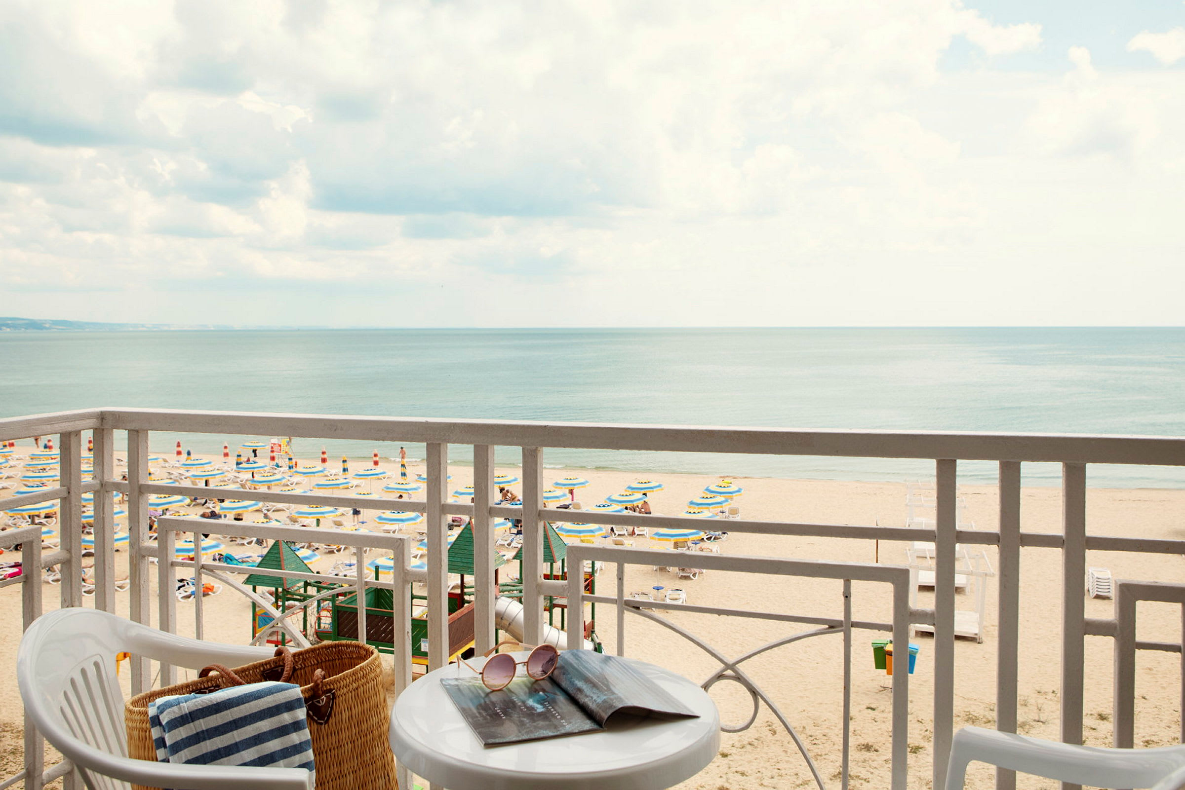 Udsigt fra balkon mod en strand med parasoller og solsenge samt havet i baggrunden. (AI-genereret tekst)