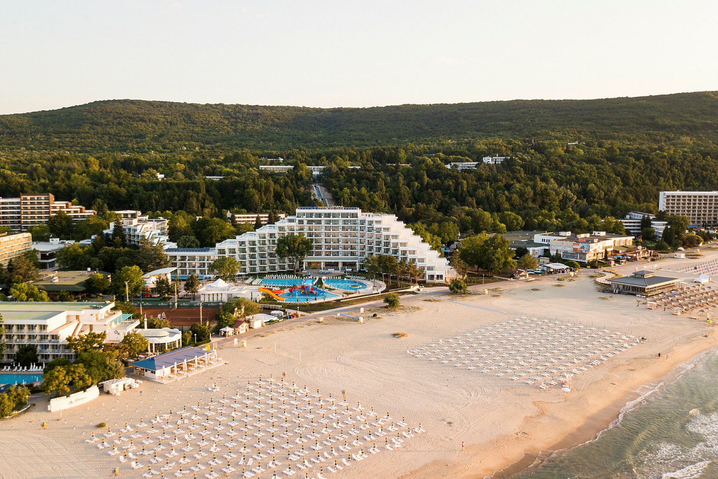Billede af en kystnær feriedestination med hotel, poolområde og strand med parasoller. (AI-genereret tekst)