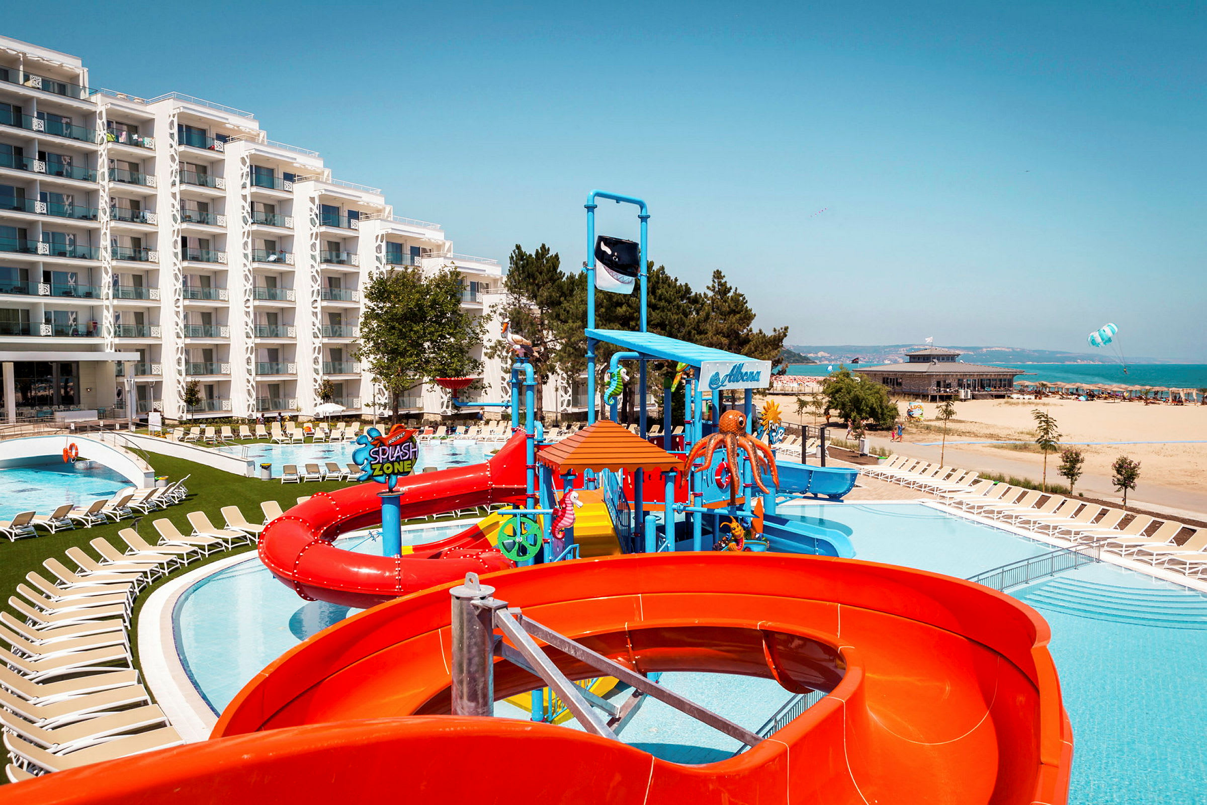 Billede af et hotelområde med pool, vandrutsjebaner og liggestole tæt på stranden. (AI-genereret tekst)