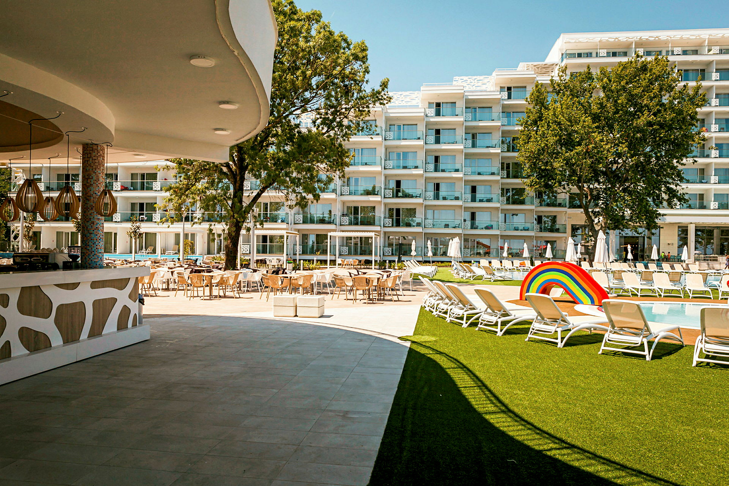 Hotelområde med pool, liggestole, græsplæne og bygninger i baggrunden. (AI-genereret tekst)