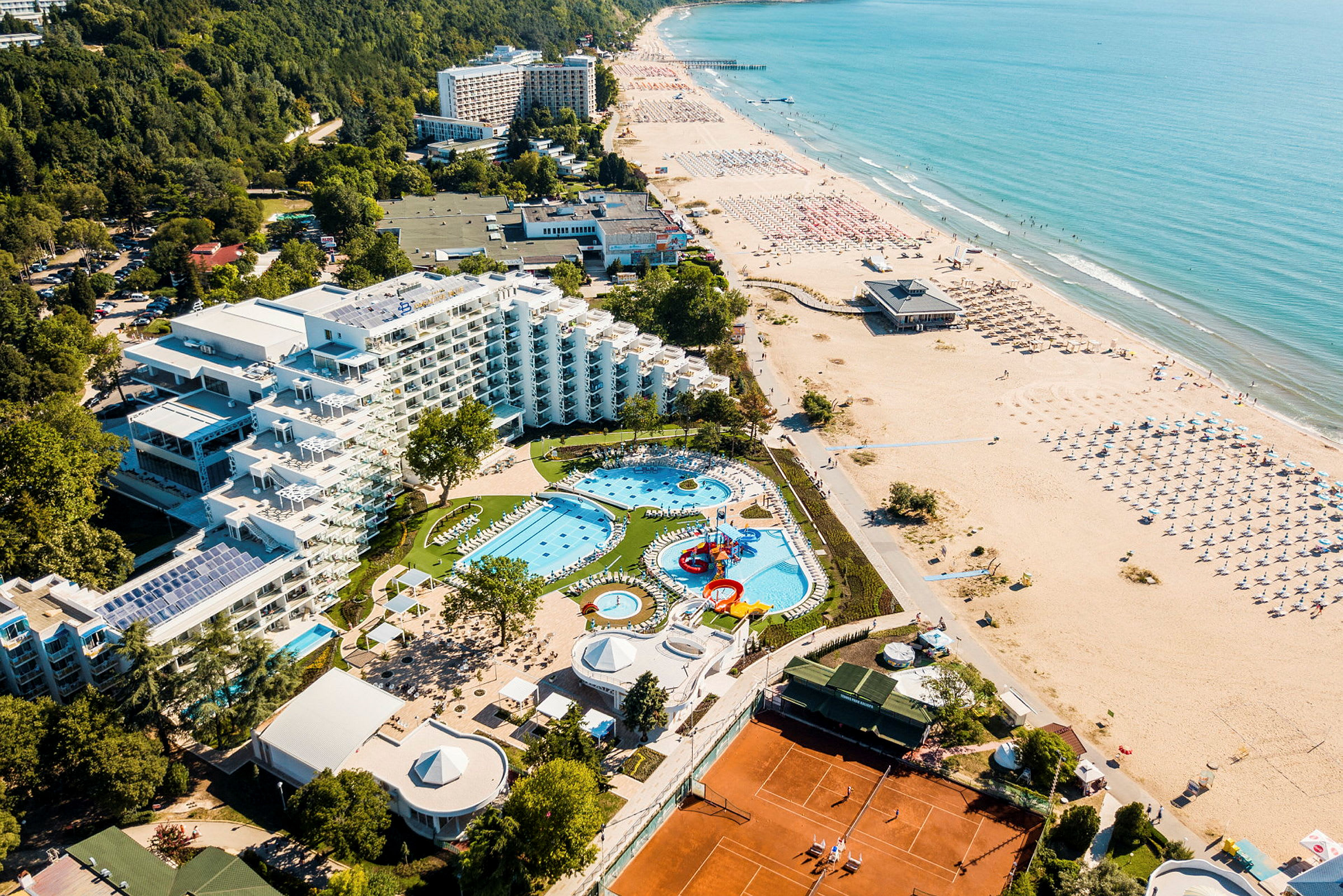 Luftfoto af hotel med poolområde, grønne omgivelser og strand. (AI-genereret tekst)