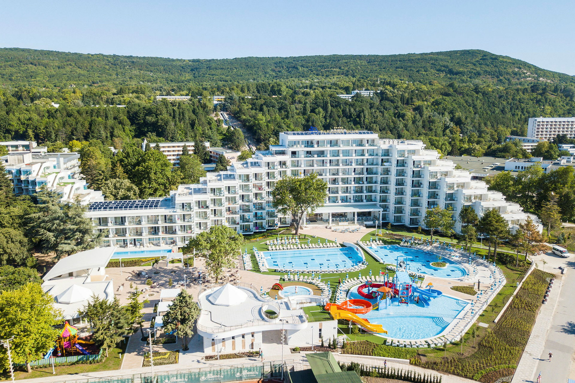 Luftfoto af et hotel med pools og legepladser, omgivet af grøntområder og mindre bygninger. (AI-genereret tekst)