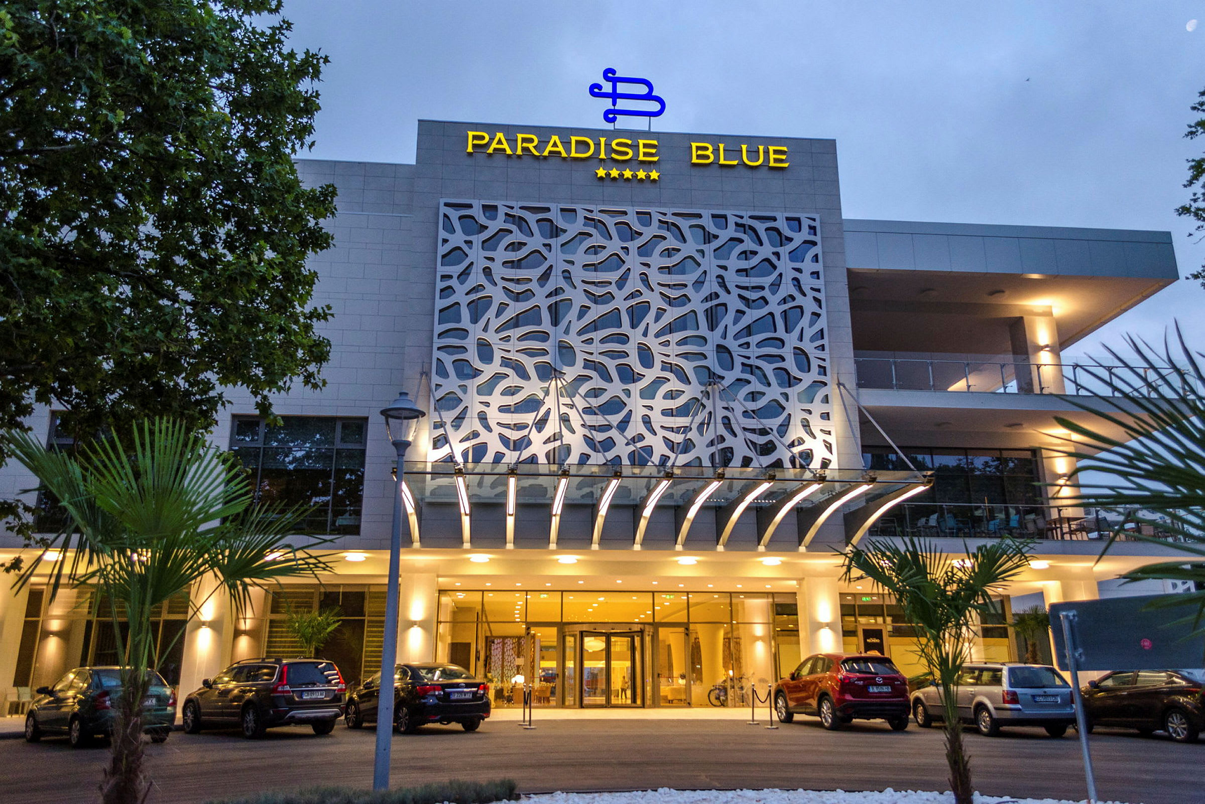 Facaden på Paradise Blue hotel med parkering og indgang. (AI-genereret tekst)