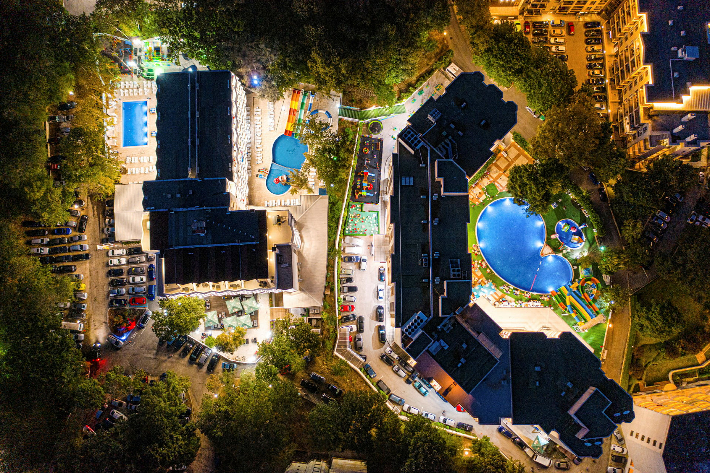 Luftfoto af hotelområde med pools, bygninger og parkeringspladser, oplyst om aftenen. (AI-genereret tekst)