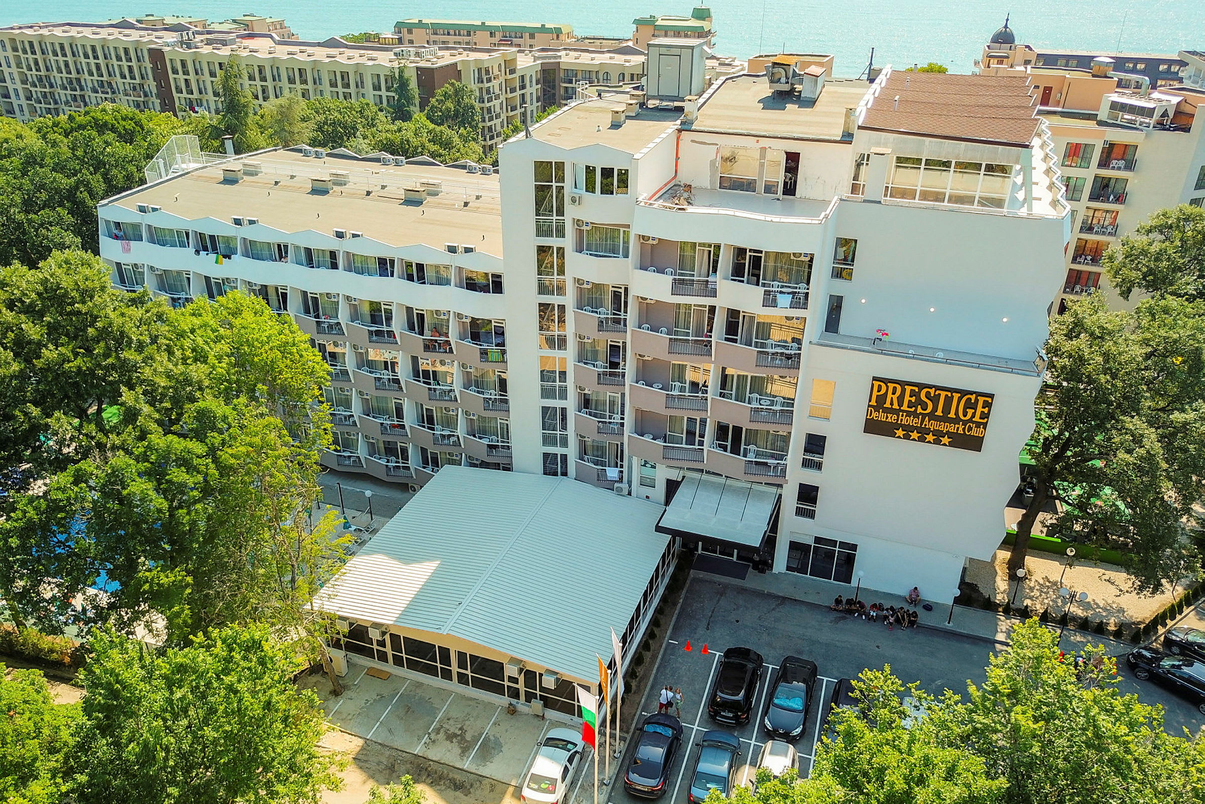 Billede af et hotelkompleks med lejlighedsbygninger og altaner, omgivet af grøntområder og med parkerede biler foran indgangen. (AI-genereret tekst)
