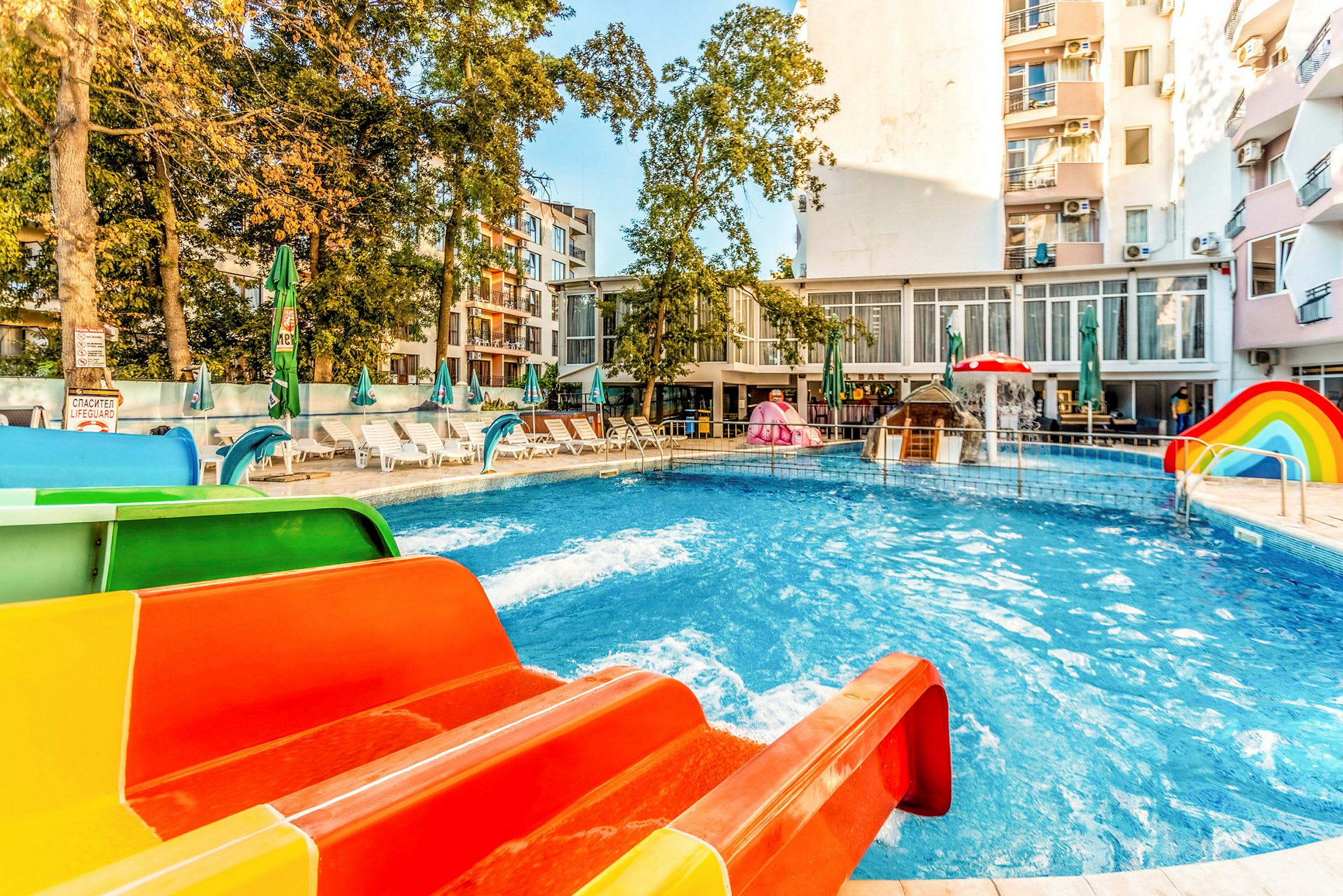 En swimmingpool med farverige vandrutsjebaner og liggestole rundt om den, omgivet af bygninger og træer. (AI-genereret tekst)