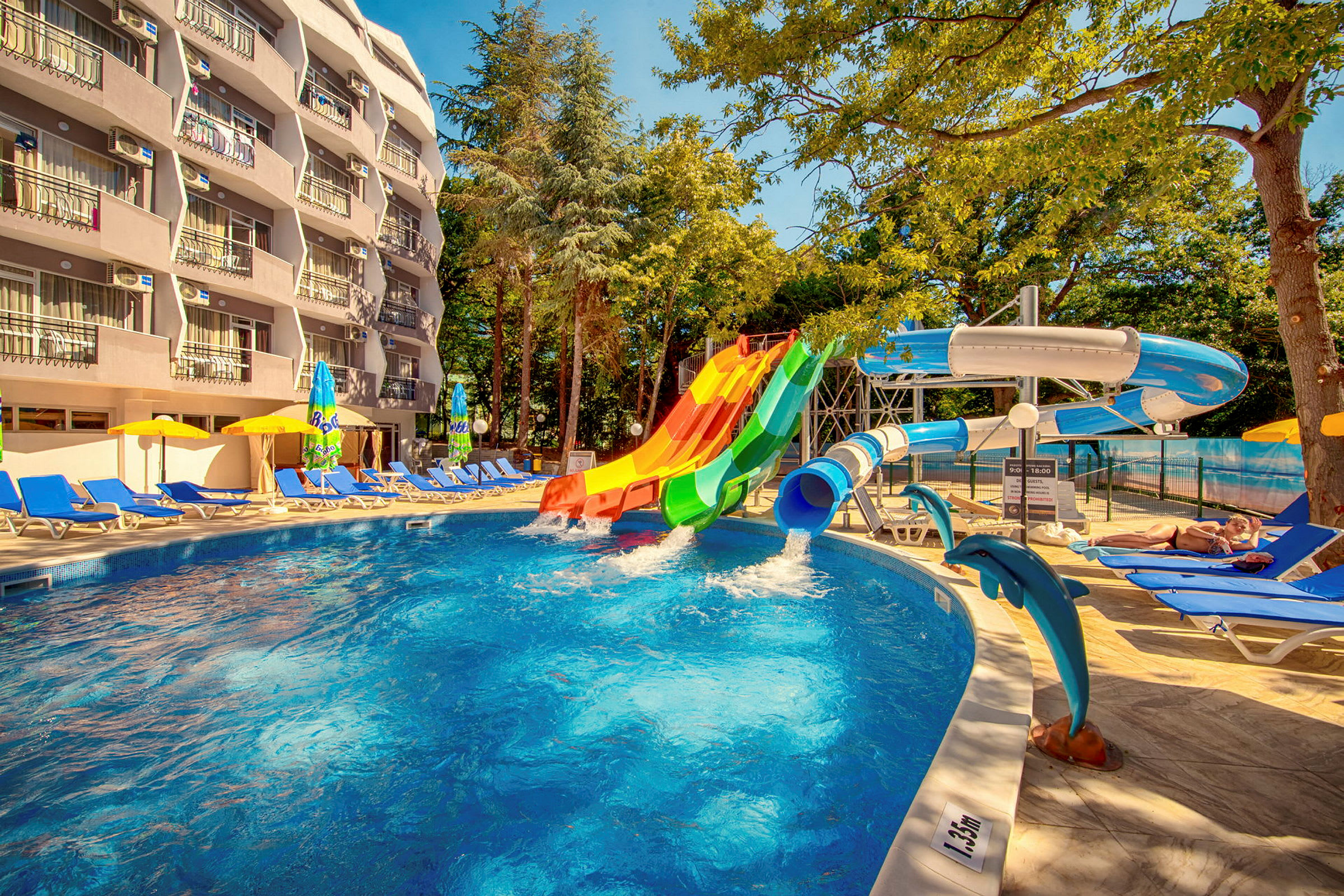 En udendørs pool med farverige vandrutsjebaner og liggestole rundt om. Et hotel med altaner er synligt i baggrunden. (AI-genereret tekst)