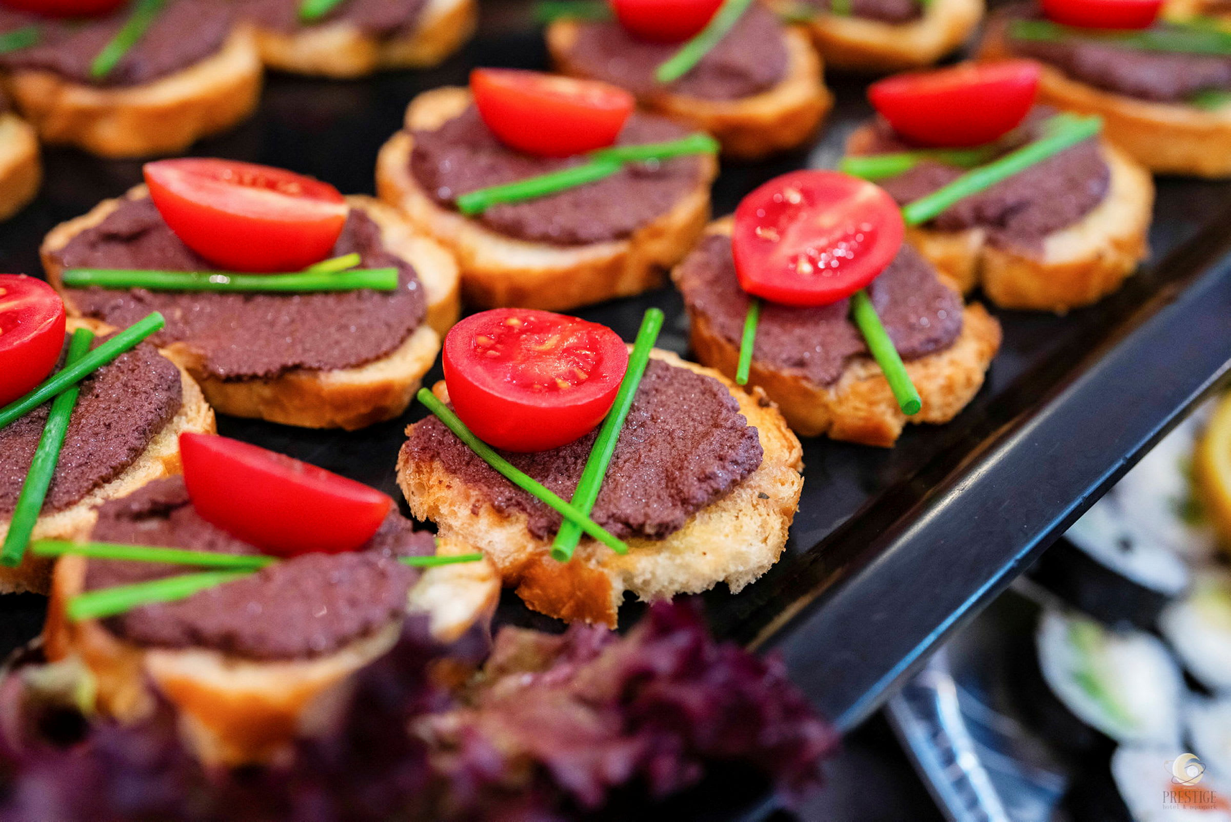 Små skiver sprødt brød med et lag mørk puré, pyntet med halve cherrytomater og stilke af purløg. (AI-genereret tekst)