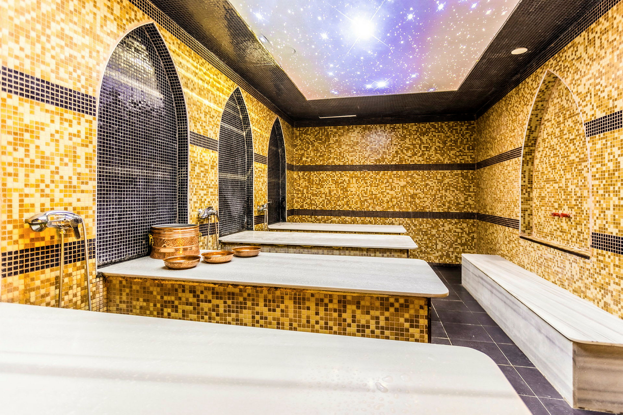 Billede af et hamam med mosaikbeklædte vægge, hvide marmorbænke og kobberskåle. (AI-genereret tekst)