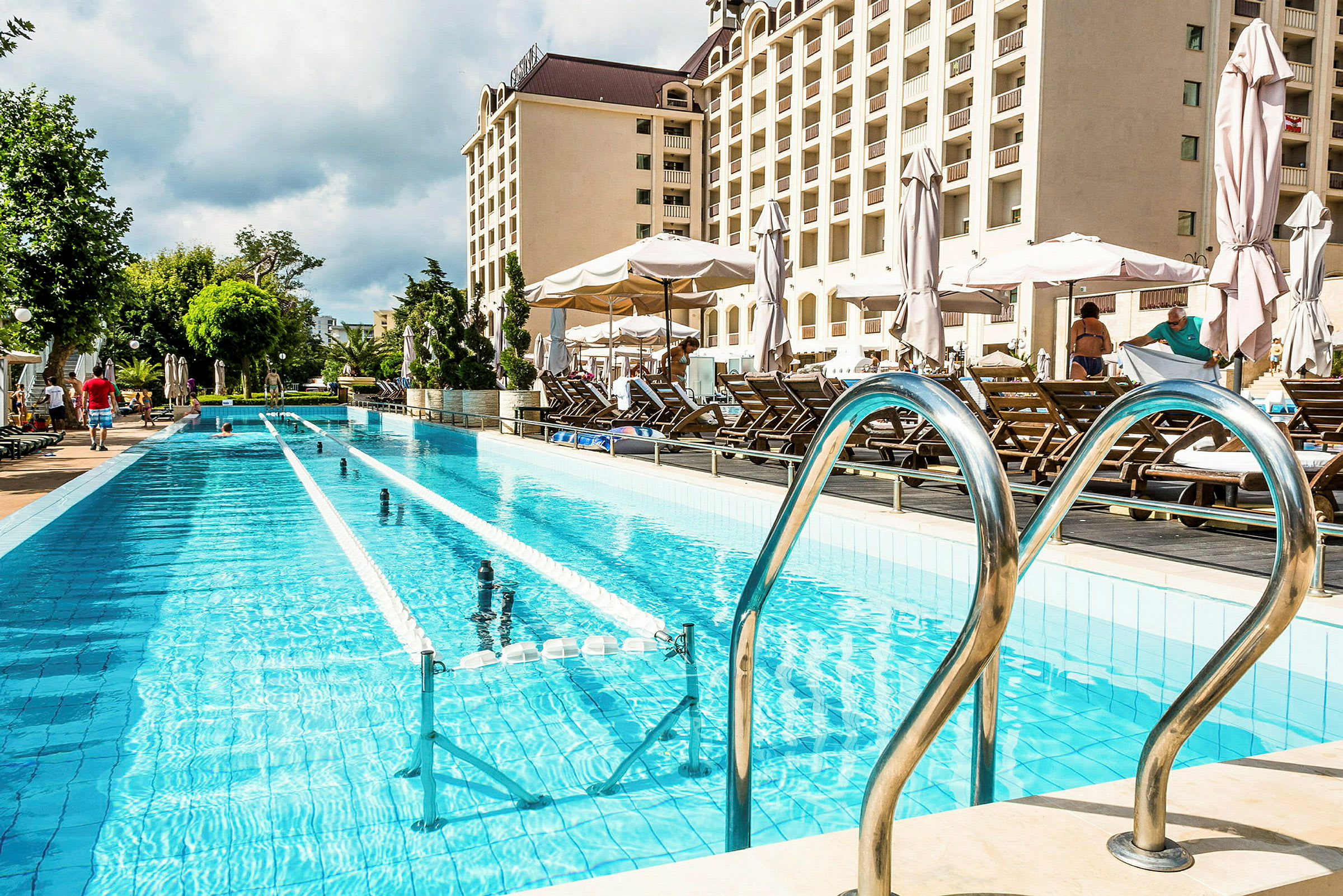 Lang swimmingpool med liggestole og parasoller ved siden af, hotelbygning i baggrunden. (AI-genereret tekst)