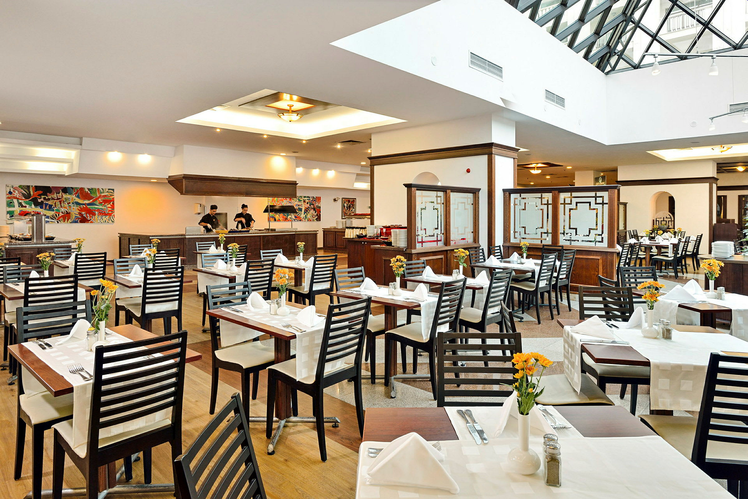 Interiør af en restaurant med borde opstillet og dækket samt personale ved serveringsdisken. (AI-genereret tekst)