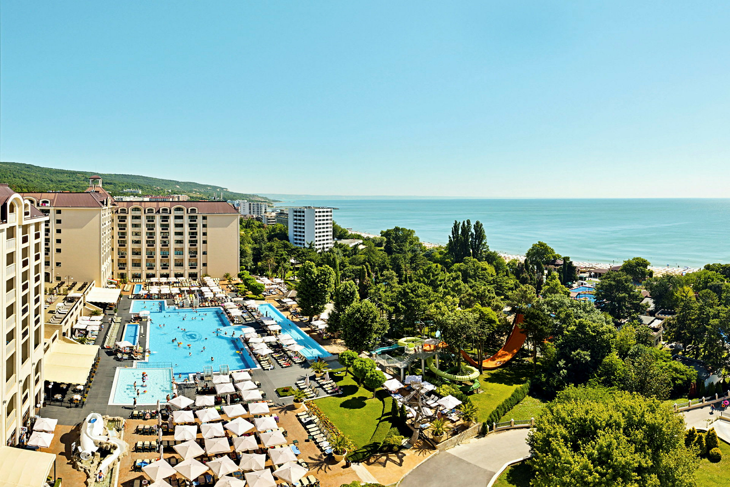 Luftfoto af hotelkompleks med pools, liggestole og grønne områder, beliggende tæt på havet. (AI-genereret tekst)