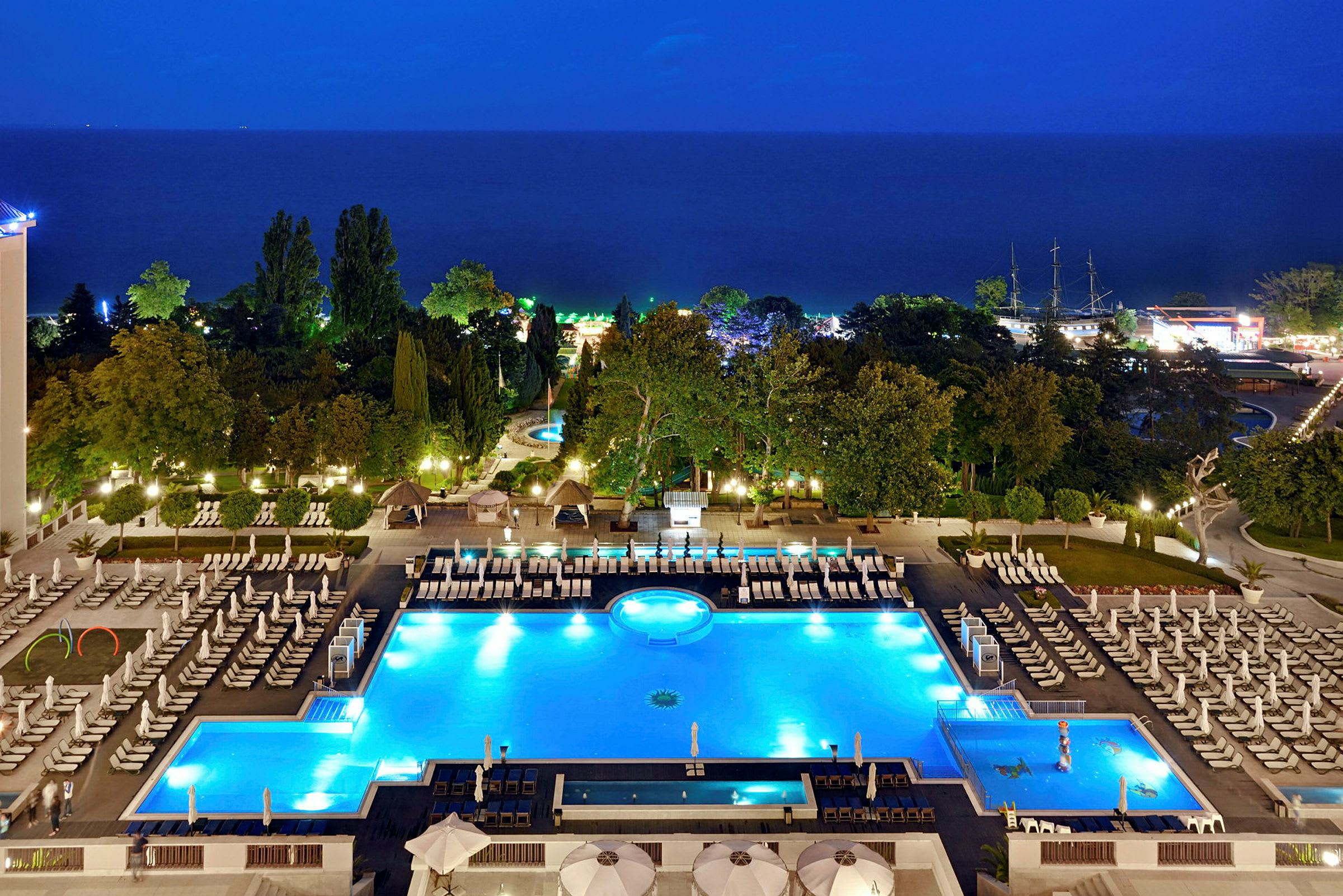 Hotelområde med oplyste swimmingpools og liggestole om aftenen, med træer og hav i baggrunden. (AI-genereret tekst)