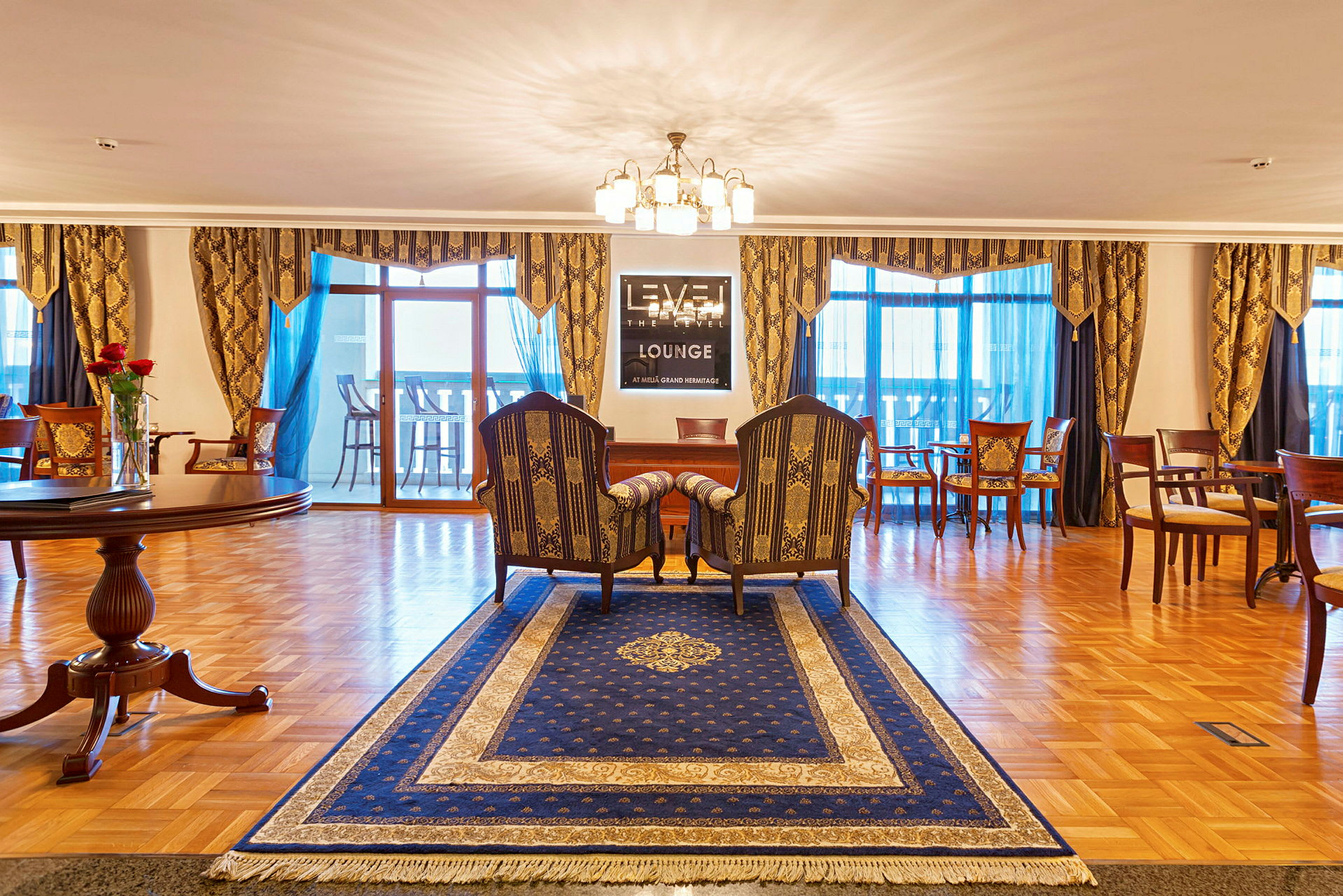 En velindrettet lounge med et blåt tæppe, et par stole og et bord, lamper og udsmykkede gardiner. (AI-genereret tekst)