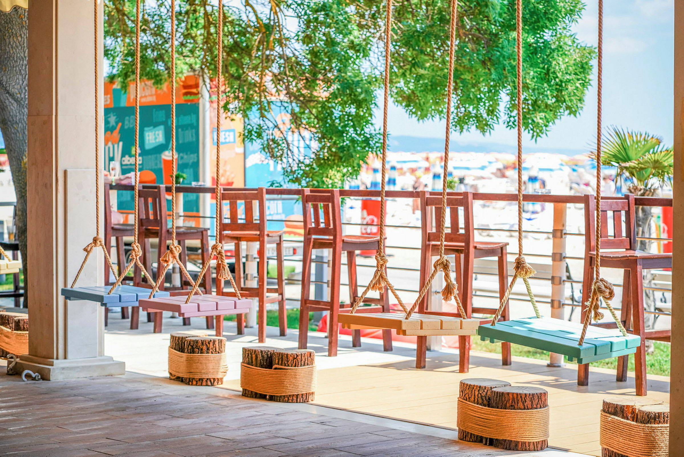 Farverige gyngesæder hænger fra reb på en terrasse med udsigt over stranden og solsenge. (AI-genereret tekst)