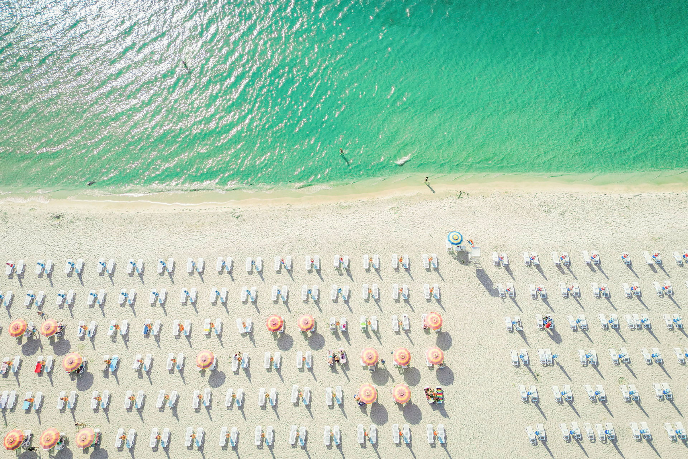 Luftfoto af en strand med parasoller og liggestole. Havet er flot grønblåt. (AI-genereret tekst)