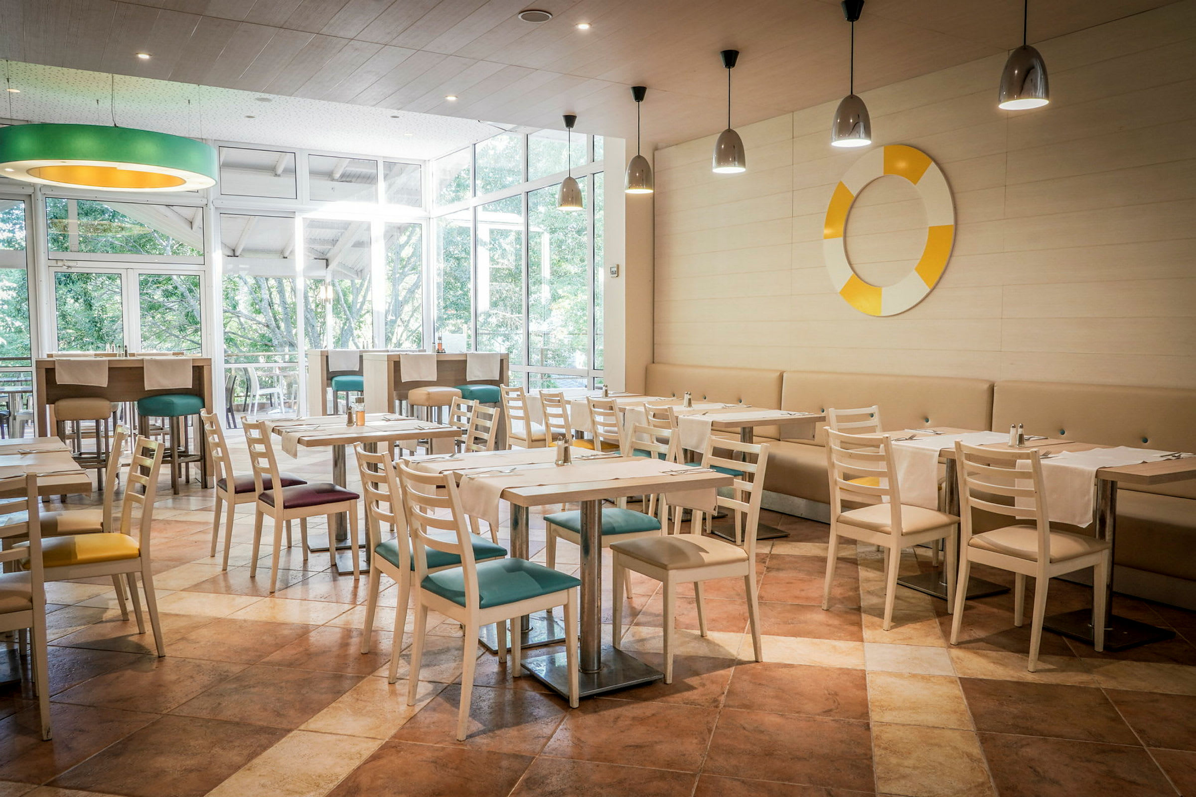 En restaurant med lyse træstole og borde, farverige siddepuder og vinduer mod grønne områder. (AI-genereret tekst)