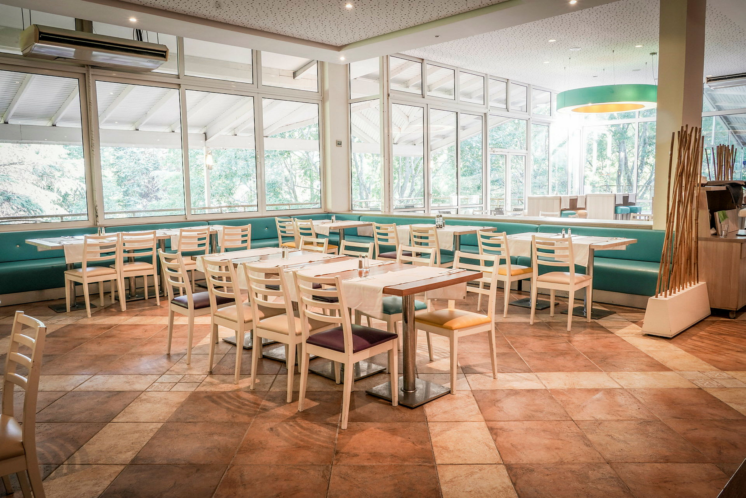 Et lyst restaurantområde med borde og stole samt store vinduer som vender mod grønt områder. (AI-genereret tekst)