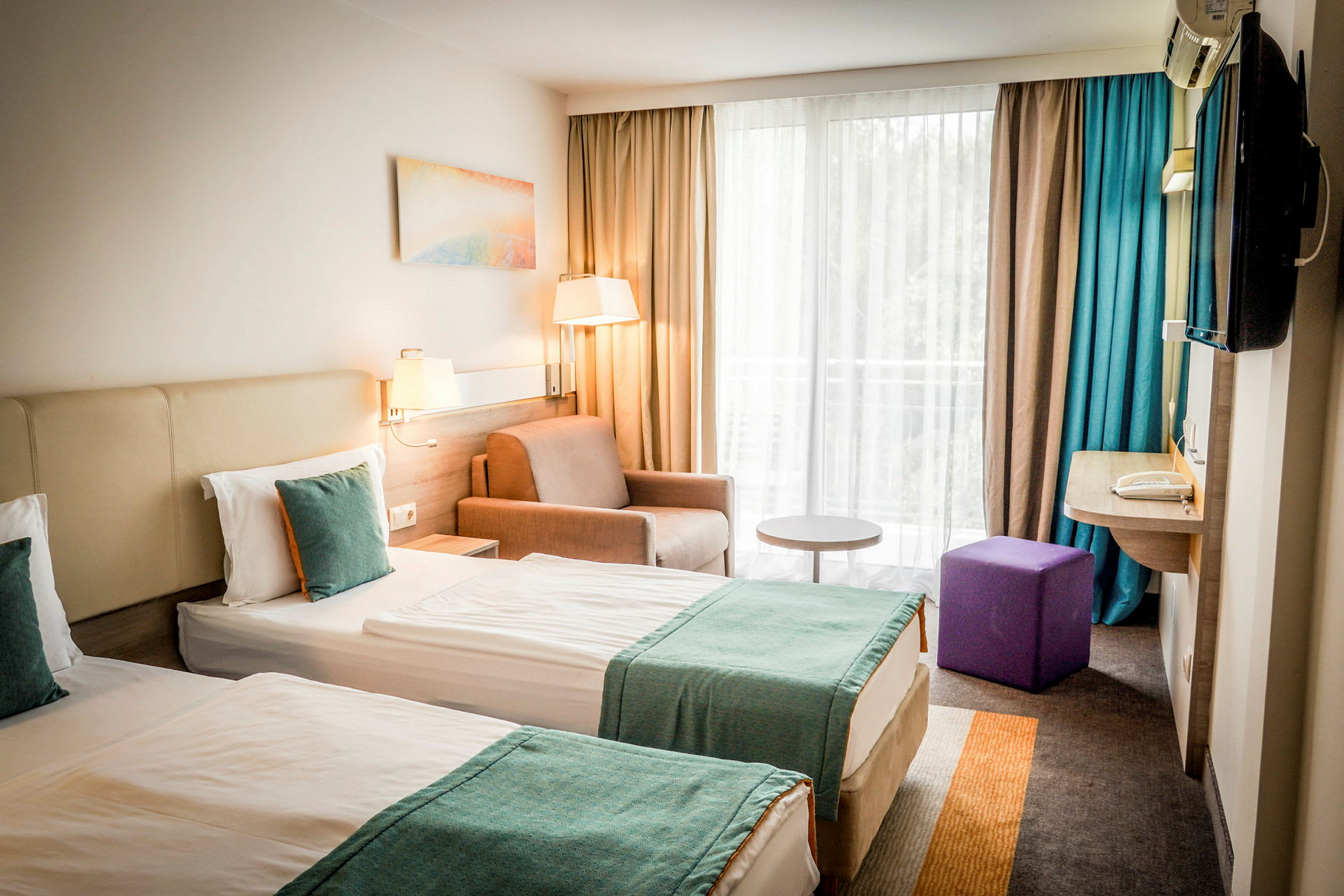 To enkeltsenge med turkise tæpper i et hotelværelse med vindue og siddeområde. (AI-genereret tekst)