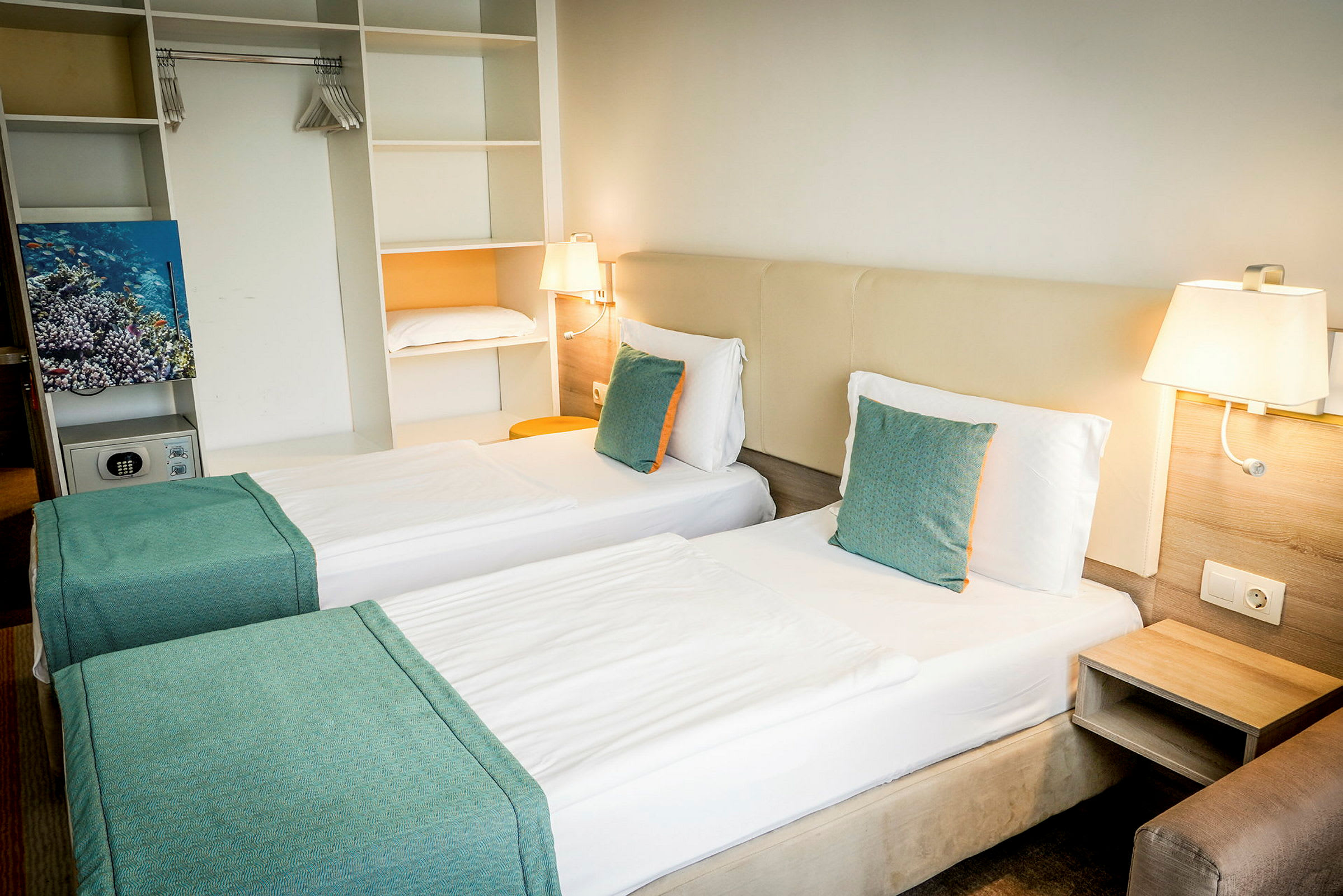 Hotelværelse med to enkeltsenge, hvide lagner og turkise puder. Indbygget opbevaring og væglamper ses. (AI-genereret tekst)