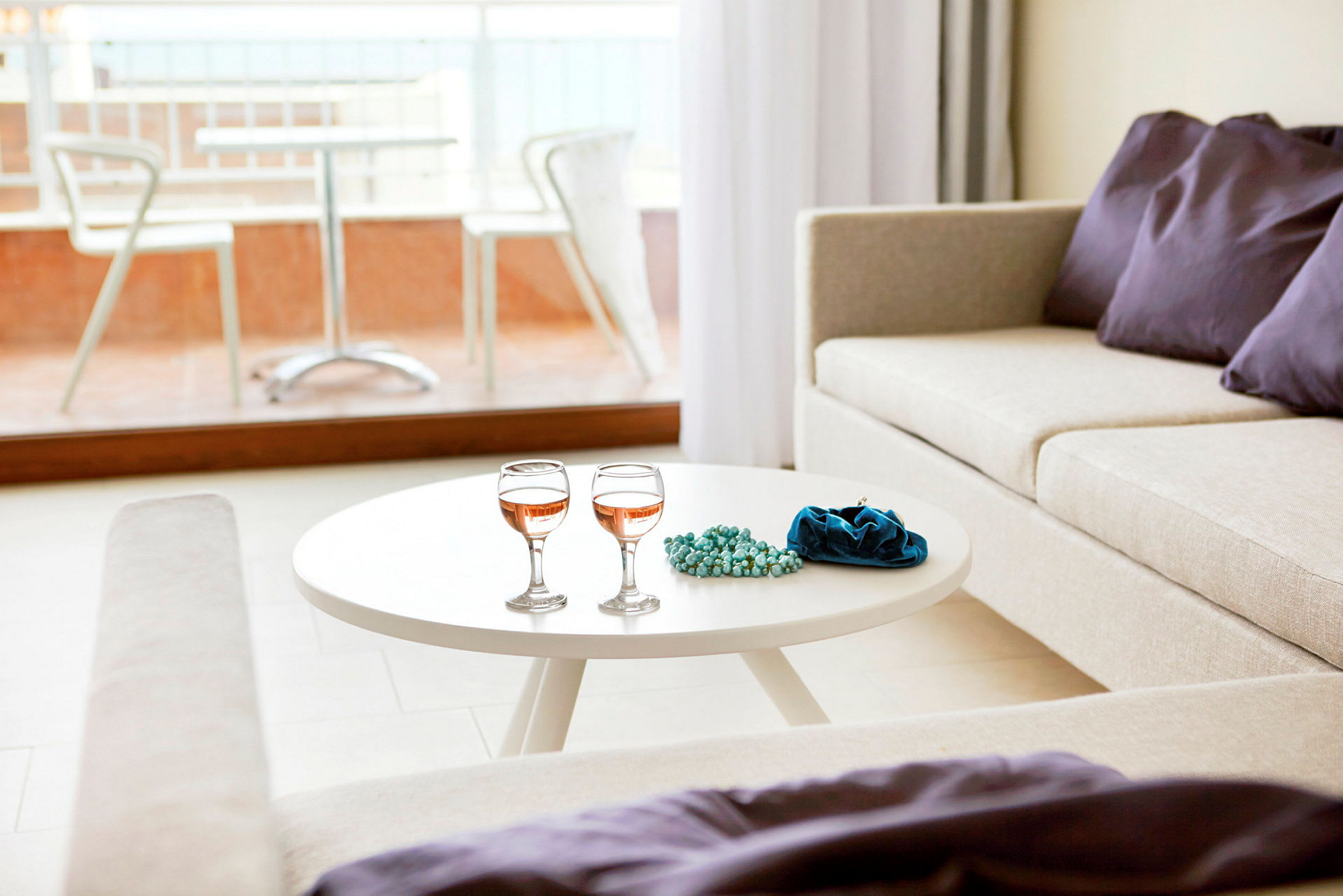 En lys stue med et hvidt sofabord, der har to vinglas, et blåt smykkesæt eller pose og perler. En beige sofa med dekorative lilla puder ved siden af, og en altan med bord og stole synlig uden for glasvinduet. (AI-genereret tekst)