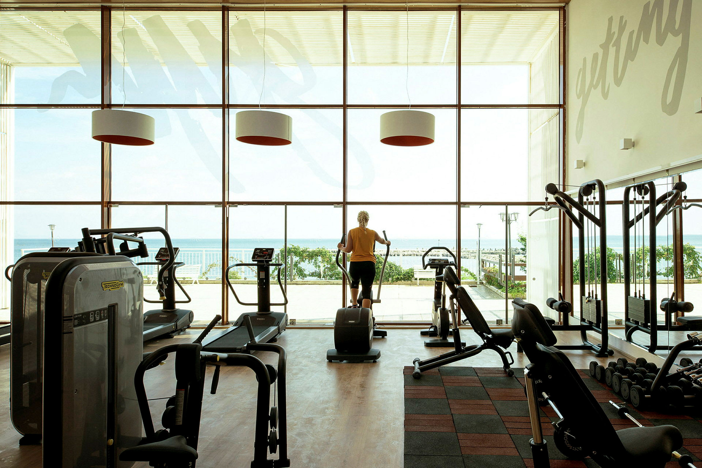 Et træningsrum med fitnessmaskiner foran store glasvinduer med udsigt over havet. (AI-genereret tekst)