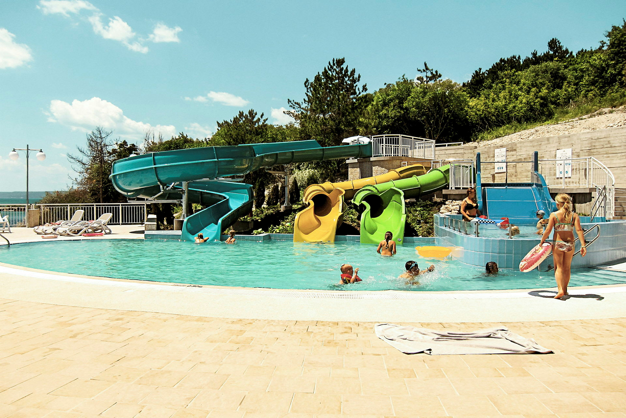 Udendørs pool med farverige vandrutsjebaner omgivet af grønne områder og liggestole. (AI-genereret tekst)
