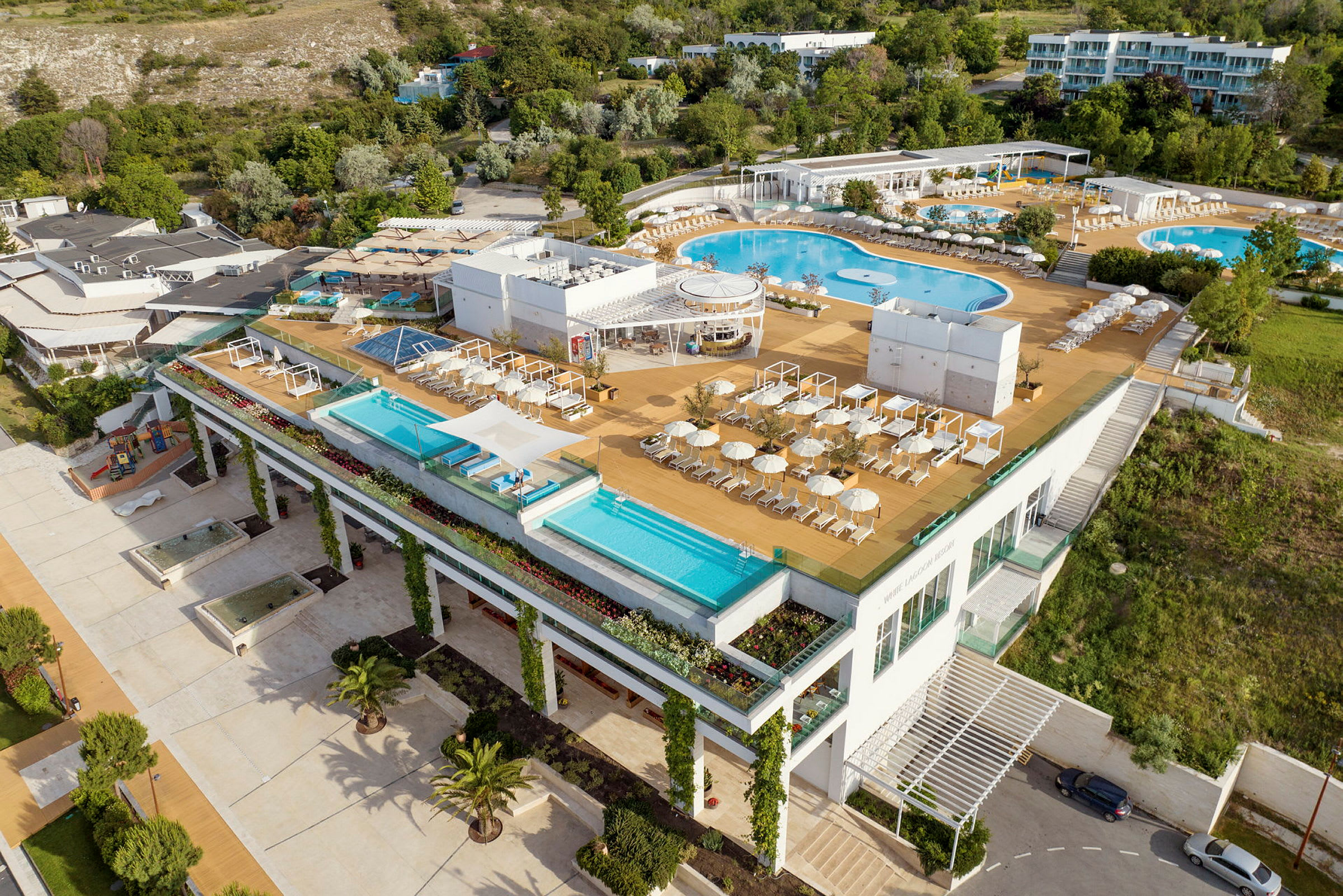 Et moderne hotelanlæg med flere udendørs swimmingpools, liggestole og parasoller, omgivet af grønne områder. (AI-genereret tekst)