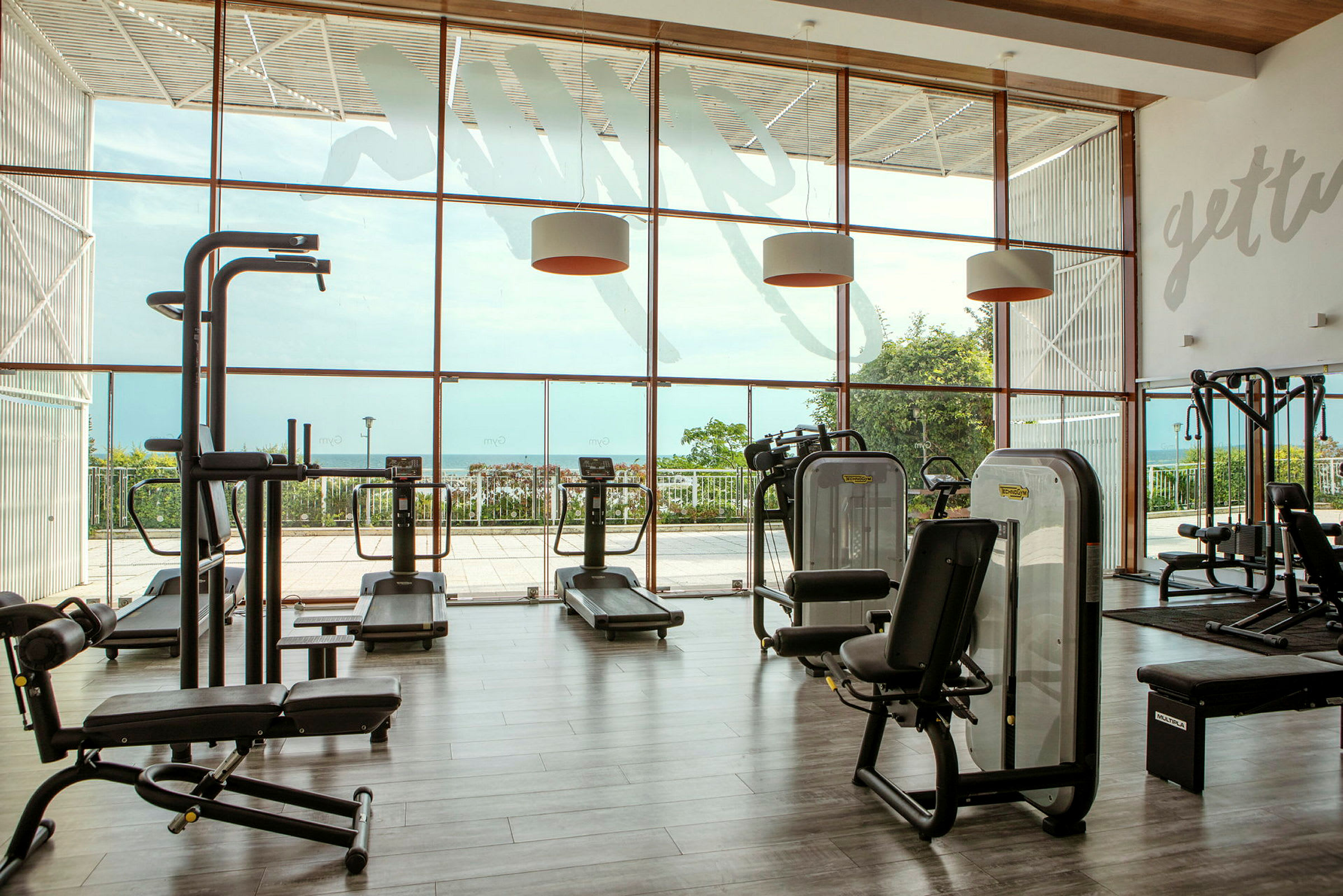 Et fitnesscenter med træningsudstyr og store vinduer med udsigt over et grønt område og havet. (AI-genereret tekst)