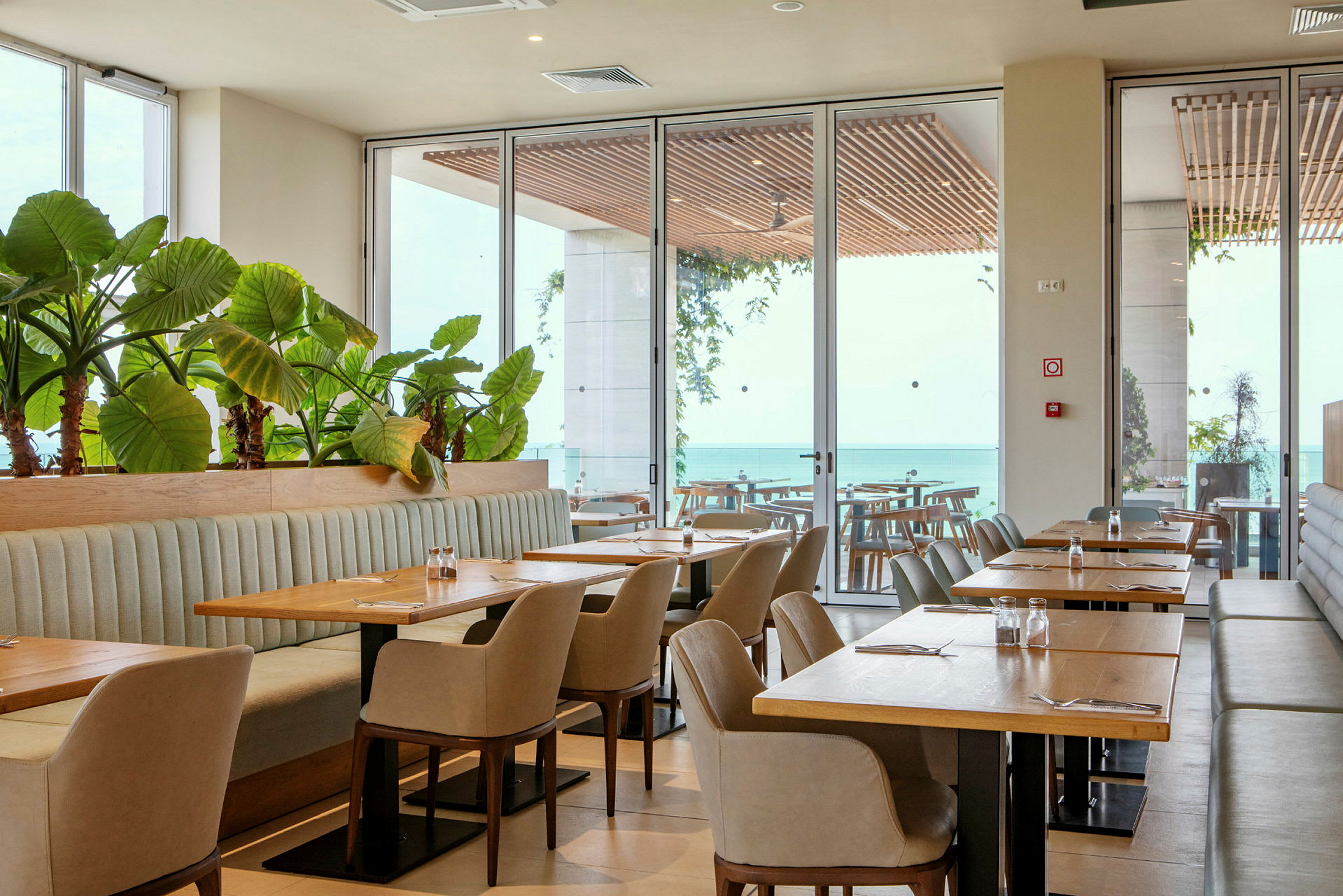 En restaurant med borde, stole og planter ved store vinduer med udsigt over havet. (AI-genereret tekst)