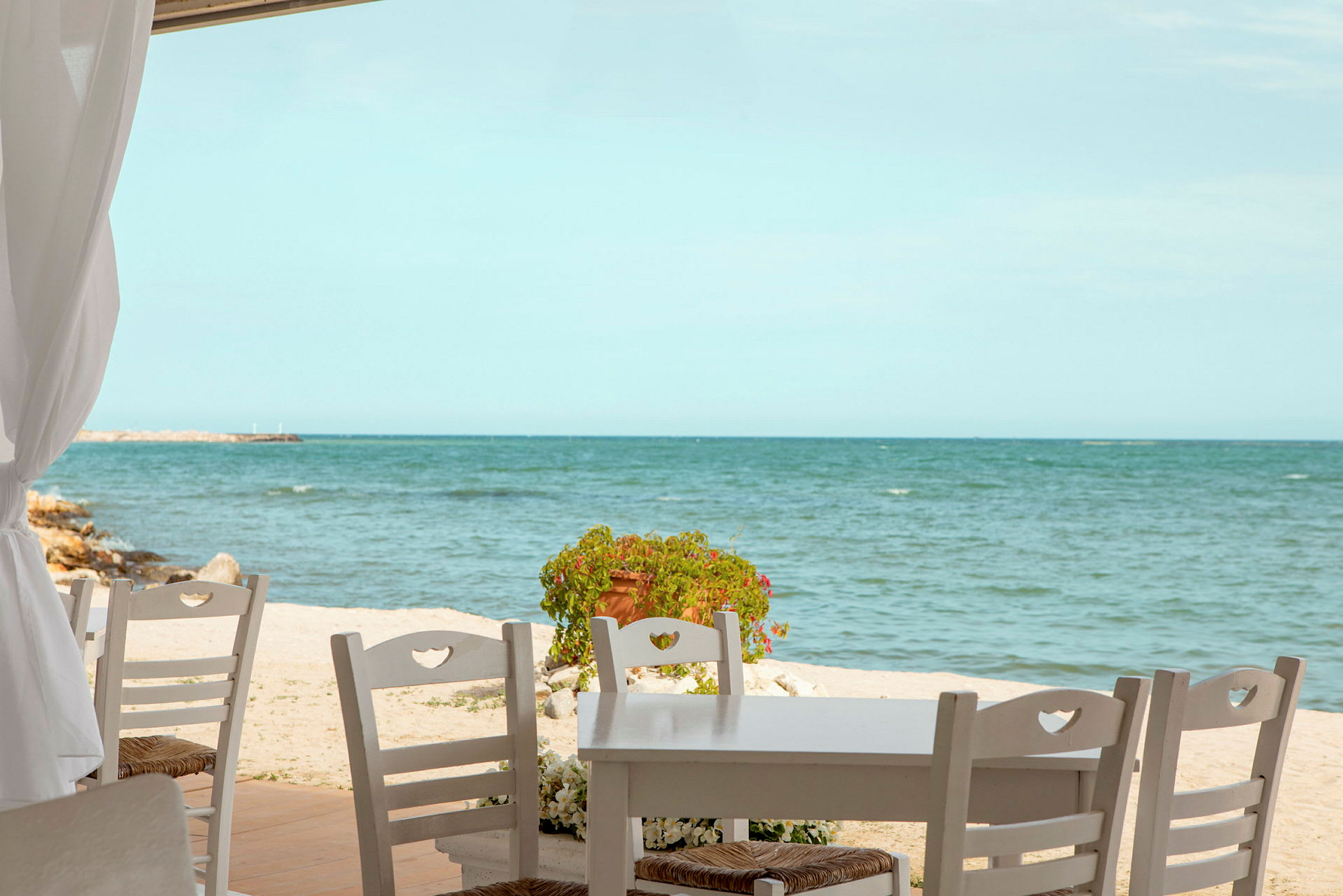 Et bord med træstole står på en terrasse tæt på en strand med havet i baggrunden. (AI-genereret tekst)