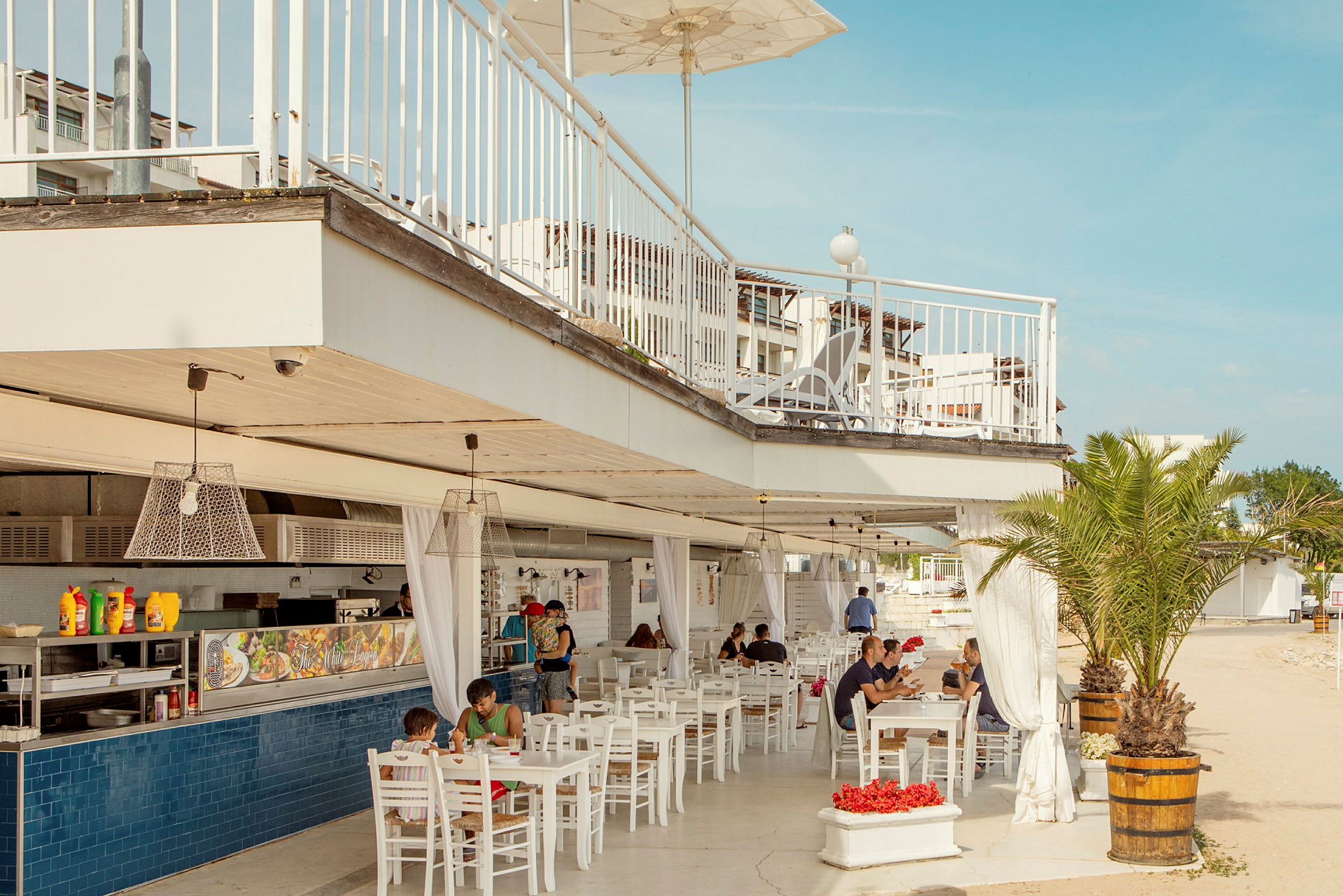 En udendørsrestaurant med hvide møbler, hængende lamper og en tagterrasse. Folk sidder og spiser under overdækningen. En palme er også synlig ved siden af restauranten. (AI-genereret tekst)