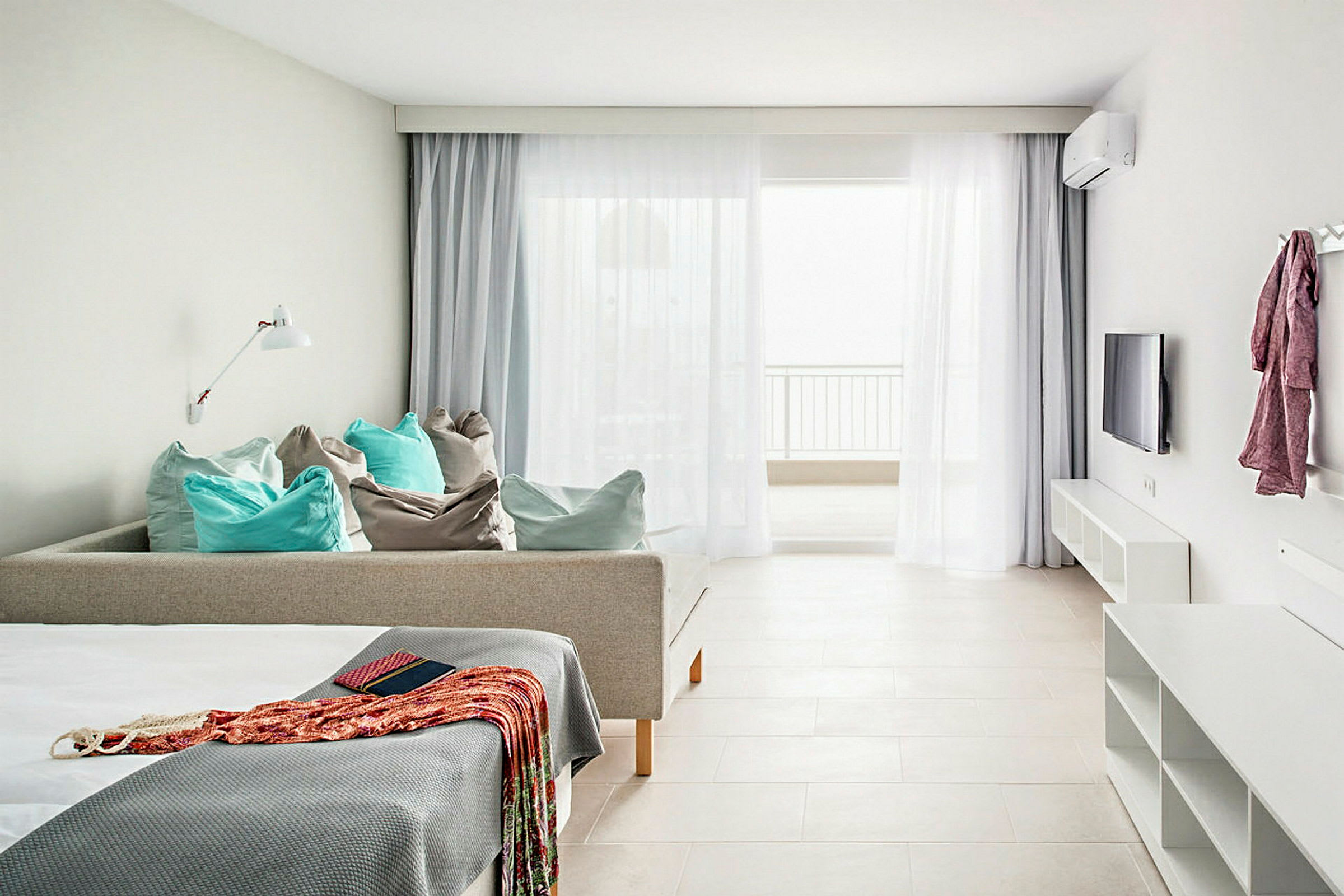 Lyst og rummeligt værelse med sofa og puder, seng med sengetæppe, vægmonteret TV og en balkon med lysindfald. (AI-genereret tekst)