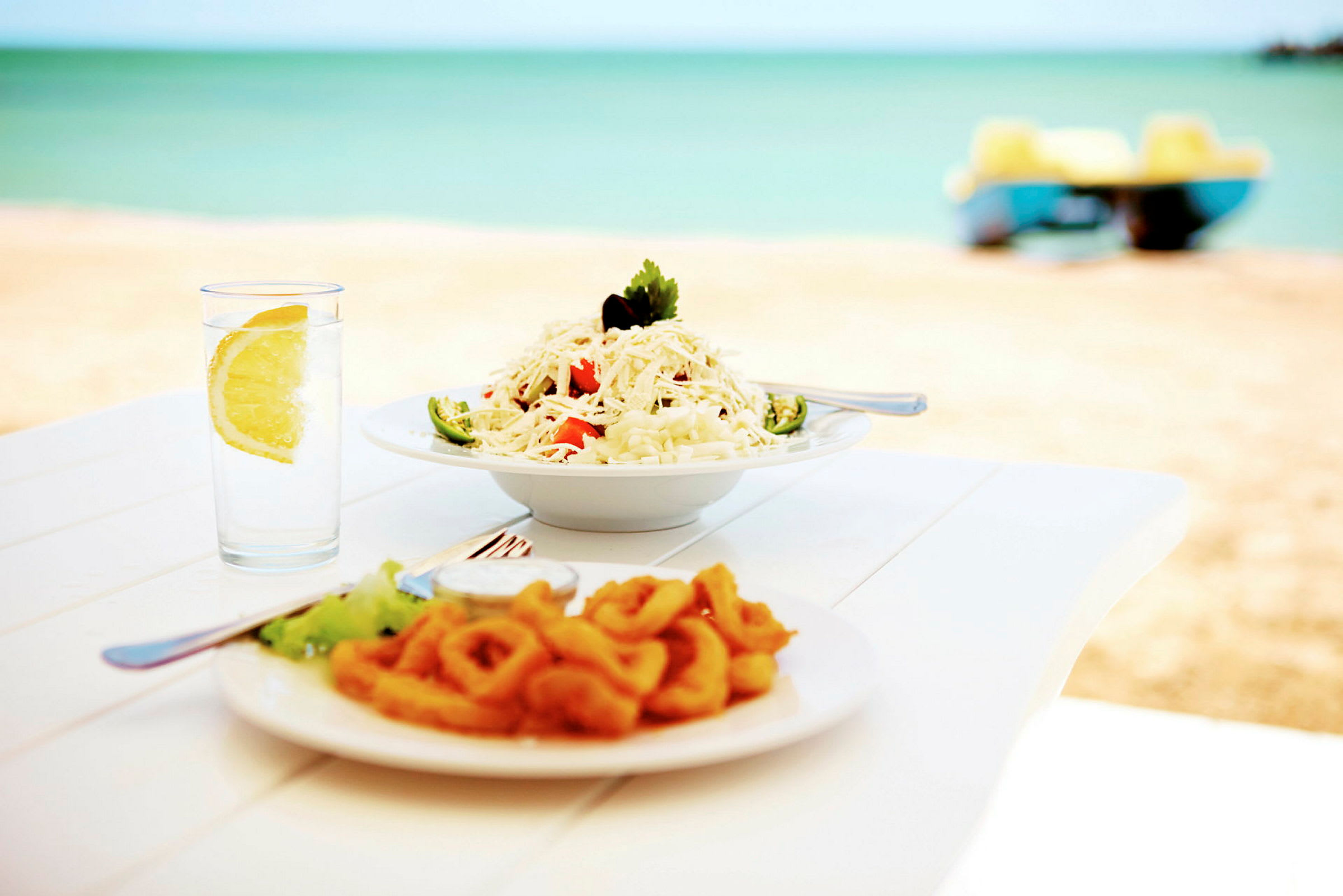 En tallerken med pasta og en tallerken med friturestegte blæksprutteringe serveret ved et bord ved stranden, med et glas vand og en citronskive ved siden af. (AI-genereret tekst)