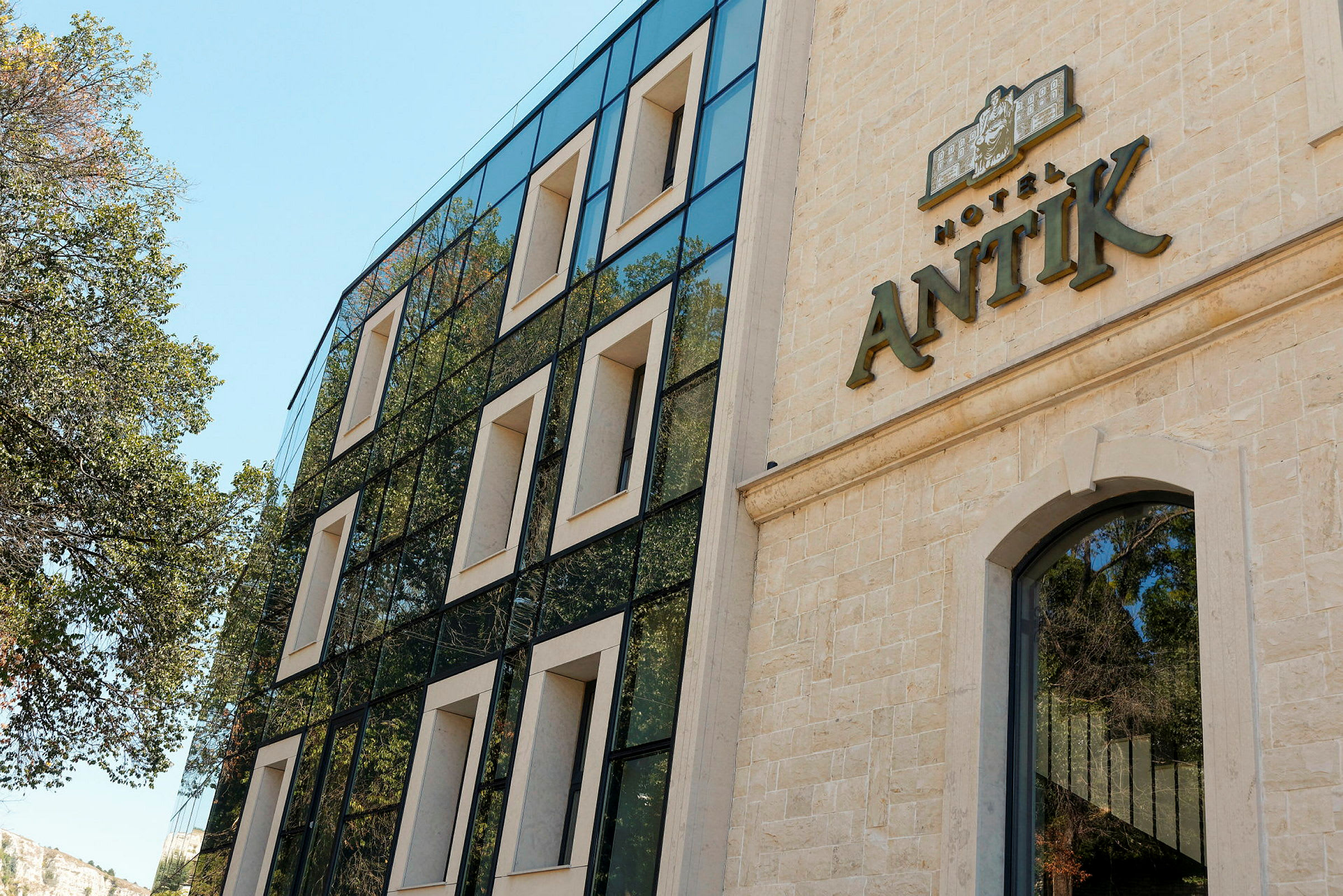 Facade af et hotel med navnet "Hotel Antik" synligt på bygningen. Den moderne bygning har store vinduer og er omgivet af træer. (AI-genereret tekst)