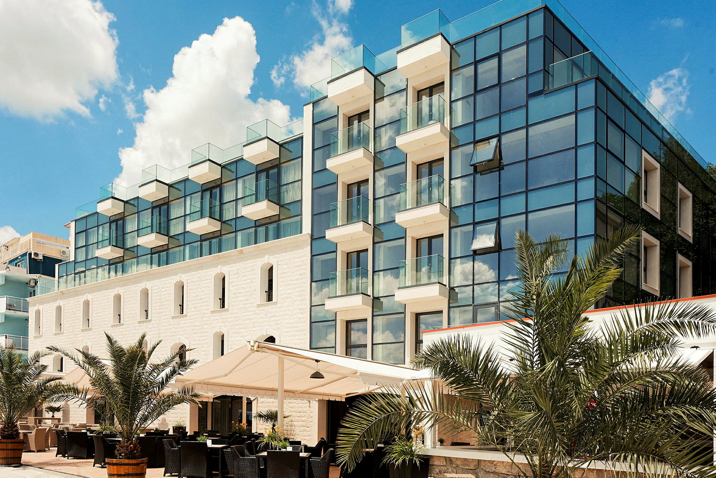 Et moderne hotel i glas og sten med altaner. Foran bygningen er der en udendørs servering med palmer. (AI-genereret tekst)