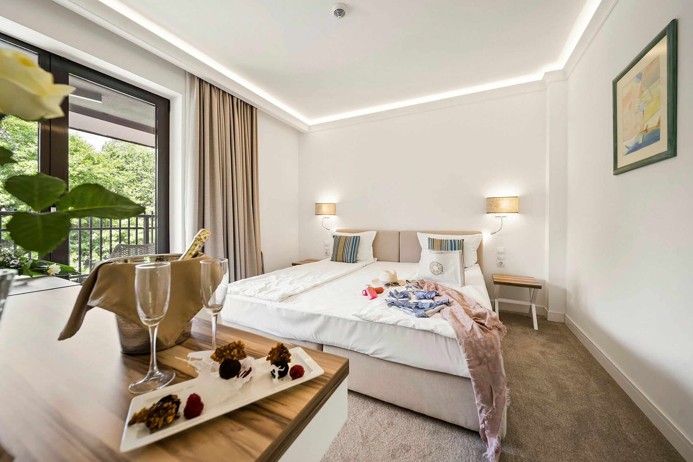 Hotelværelse med dobbeltseng, natbord og altan med udsigt til træer. Et bord med champagnekøler og dessert. (AI-genereret tekst)