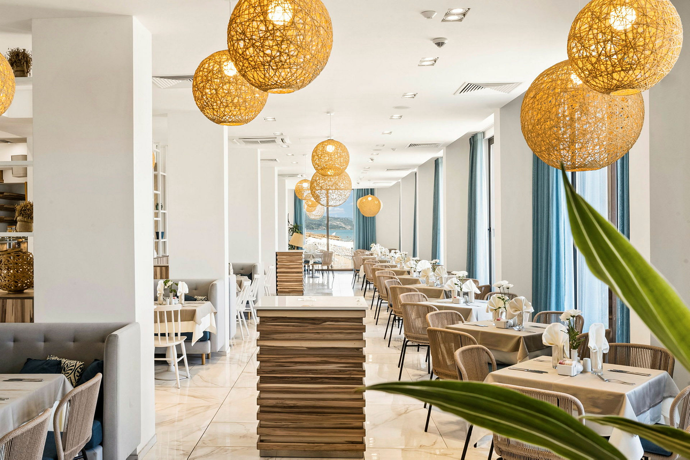 En moderne restaurant med en lys indretning med træborde, stole og store vinduer med blå gardiner. Dekorative belysningsarmaturer hænger fra loftet. (AI-genereret tekst)