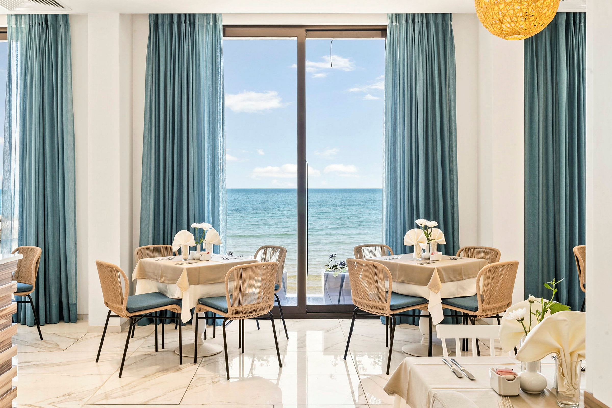 Interiør af en restaurant med borde og stole, vinduer dækket med blå gardiner, og udsigt over havet. (AI-genereret tekst)
