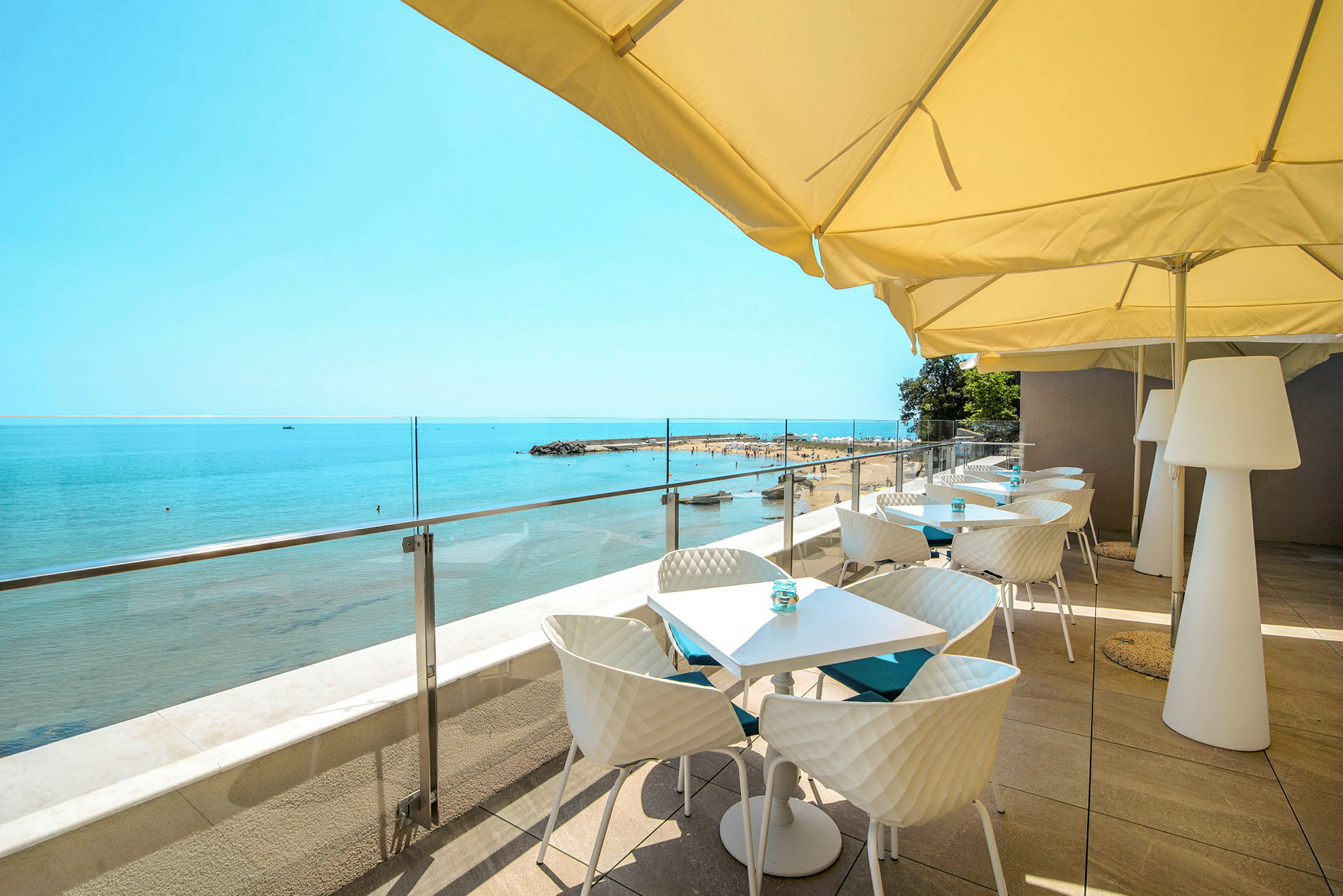 Terrasse med borde og stole under parasoller med udsigt mod stranden og havet. (AI-genereret tekst)