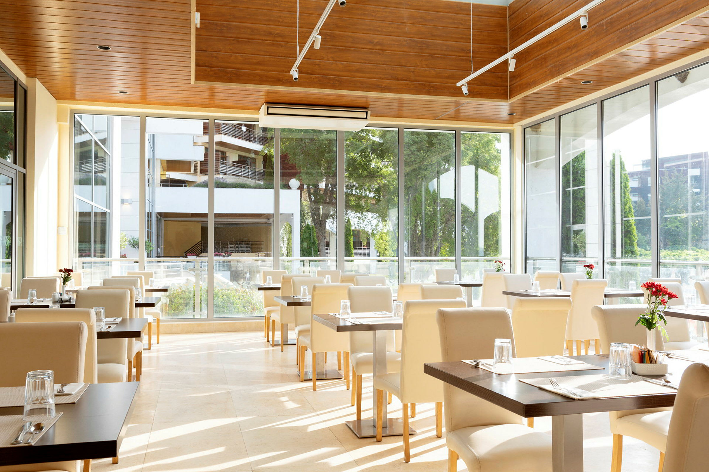 Restaurant med store vinduer, hvide stole og dækkede borde med glas og røde blomster. (AI-genereret tekst)