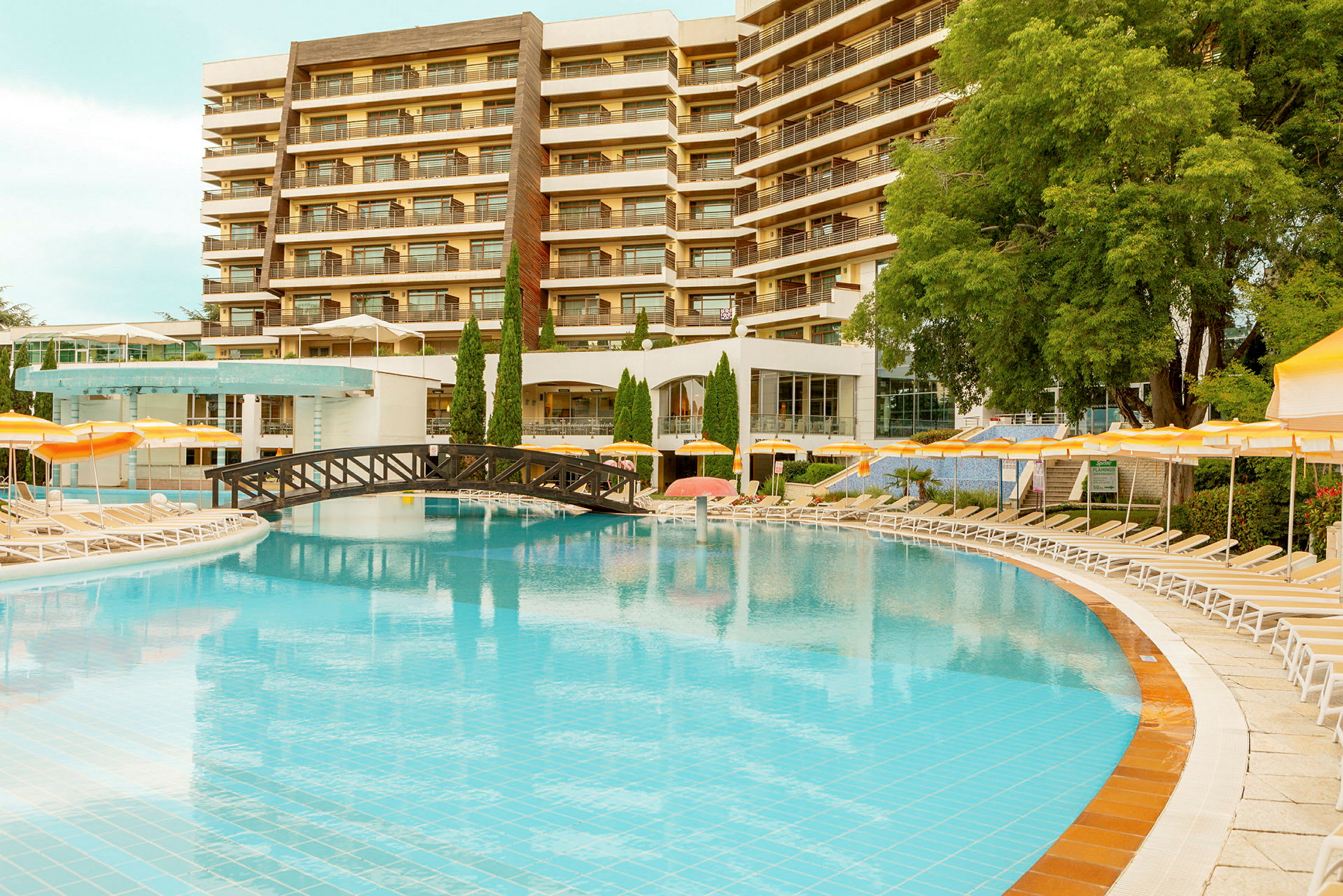 Hotelbyggning med flere etager ses i baggrunden bag en stor swimmingpool og liggestole. Balkoner og grønne omgivelser omkranser området. (AI-genereret tekst)