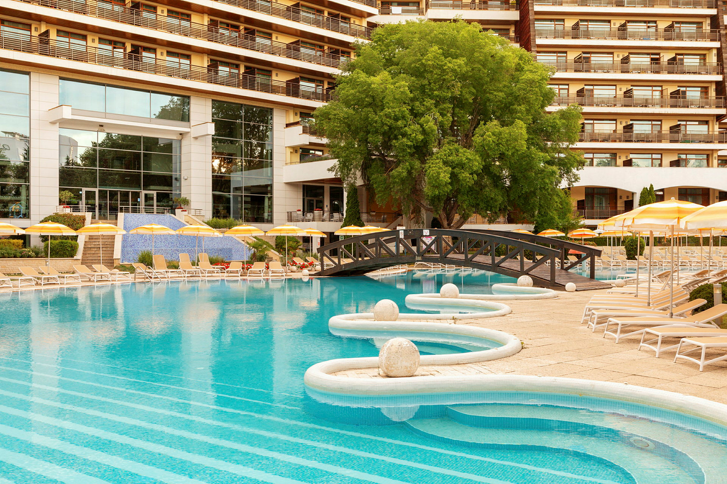 En udendørs pool med liggestole og parasoller foran et hotel. (AI-genereret tekst)