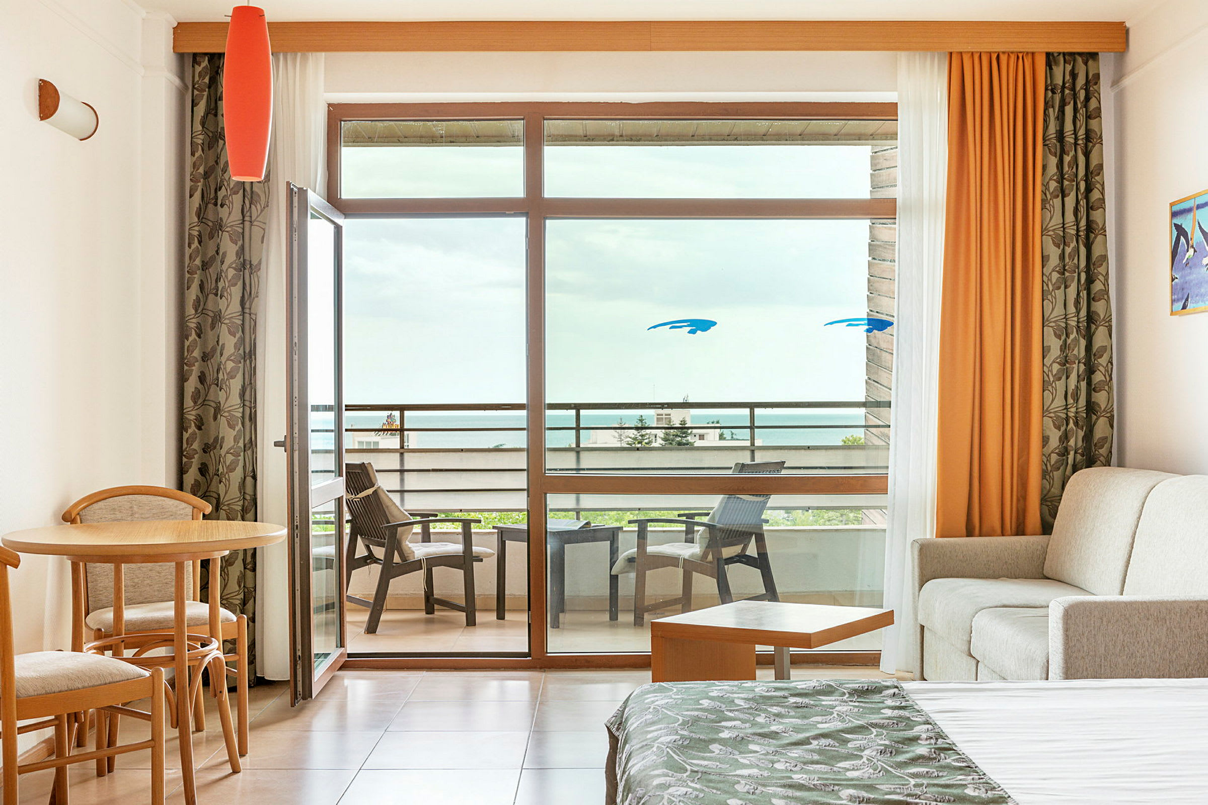 Et hotelværelse med siddeområde, seng og udsigt til en møbleret balkon mod havet. (AI-genereret tekst)