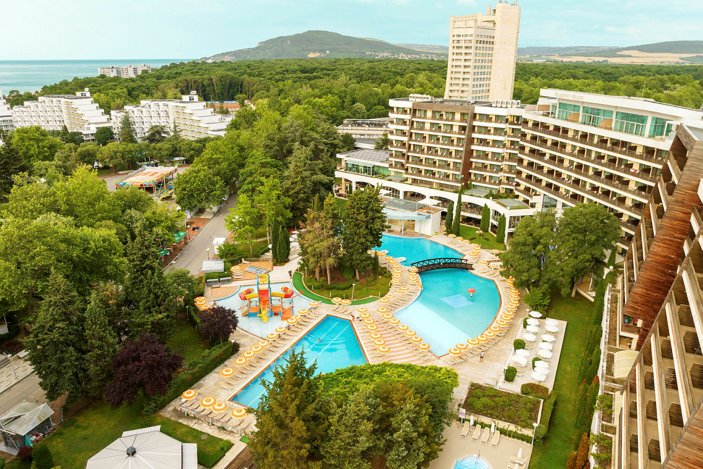 Udsigt over et hotelområde med pools, liggestole og omgivende træer. (AI-genereret tekst)